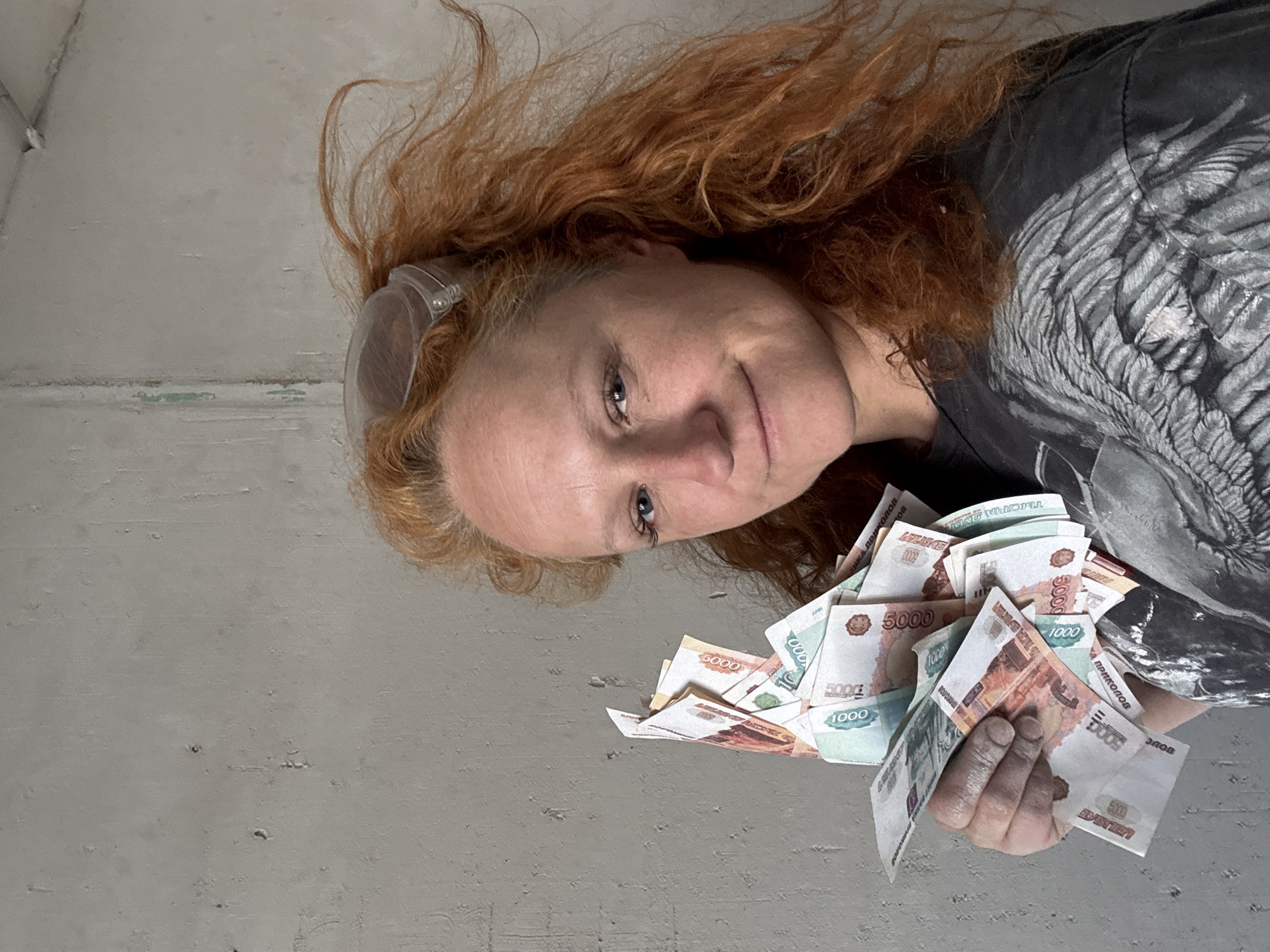 The image depicts a person with red hair, wearing safety glasses and a dark t-shirt with a grey wing design, holding a large amount of Russian rubles, specifically 5000 and 1000 denominations. The person is looking at the camera with a slight smile. The background appears to be a rough, grey concrete wall. The image composition suggests the person is lying down, viewing the camera from below. The OCR indicates the presence of “Chaikovsky, Russia” and mentions “Double,” indicating a possible reference or connection to the location and/or the value of the rubles. The hand holding the money appears slightly soiled.