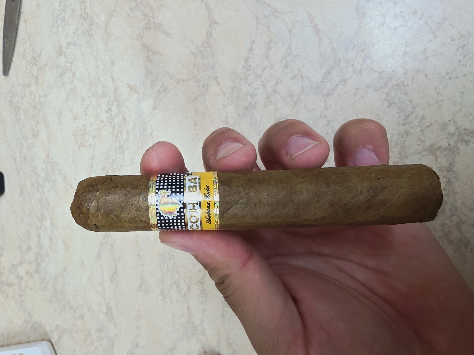 A person's hand holds a Cohiba cigar with the band clearly visible. The background is a light-colored countertop with a pattern.
