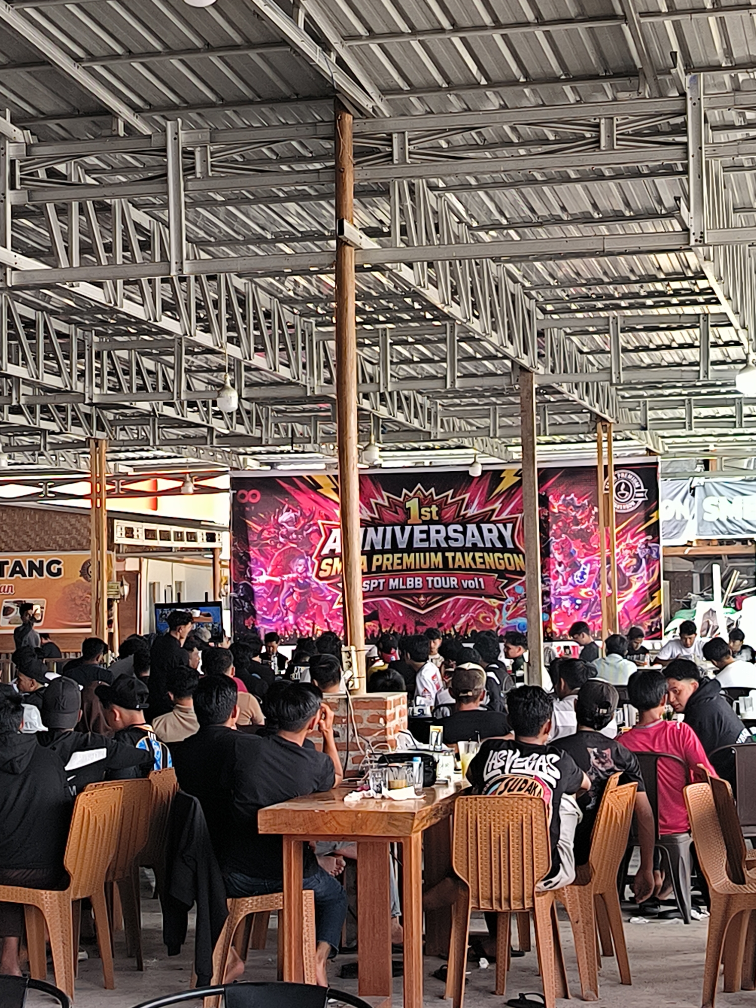 The image depicts an indoor gathering at an establishment in Nunangantara, Indonesia, likely a restaurant or event space, characterized by a prominent banner celebrating a "1st ANNIVERSARY PREMIUM TAKENGON SPT MLBB TOUR vol1". A large, colorful banner with stylized graphics and text dominates the background. The venue appears to be open-air with a corrugated metal roof supported by a metal frame structure and wooden beams. In the foreground and midground, a crowd of predominantly young men are seated at wooden tables, many of them facing away from the camera. They are dressed casually, many in black t-shirts. The tables are set with drinks and some food items. The lighting is diffuse, suggesting it's daytime. The overall atmosphere suggests a social event or watch party related to the Mobile Legends: Bang Bang (MLBB) esports tournament.