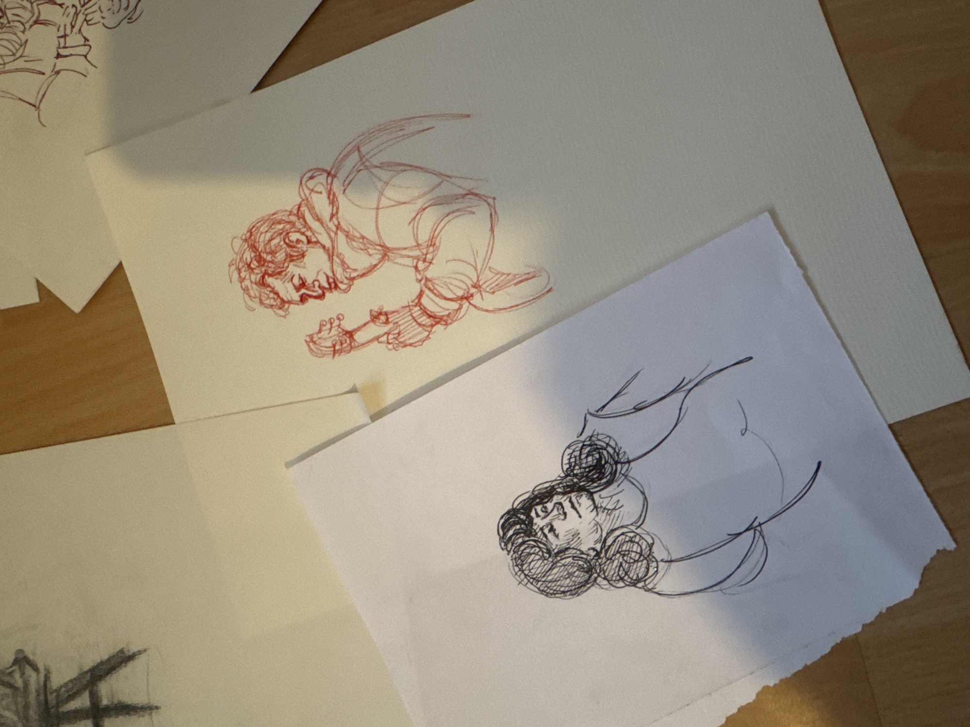 The image shows several pieces of paper scattered on a light-colored wooden surface. The papers contain sketches. One sketch, drawn in red ink, depicts a person kneeling and playing a guitar. Another sketch, in black ink, depicts a person's head with elaborate hair. The other pieces of paper have portions of sketches as well, but the details are not clear. The scene appears to be indoors. No information from the image indicates the location is in Burgas, Bulgaria.