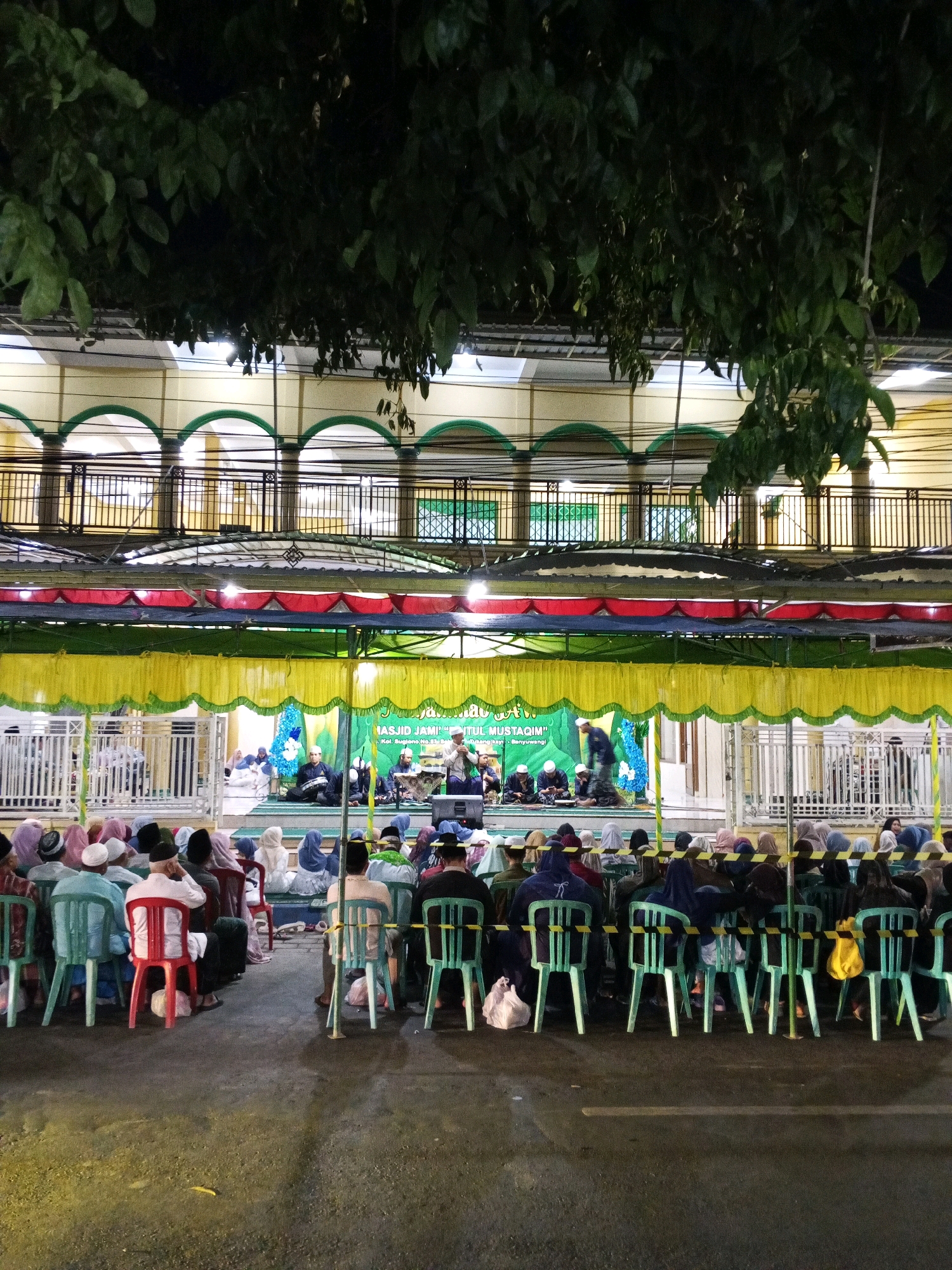 A wide-angle, nighttime, outdoor photograph captures a gathering of people seated on chairs, facing a stage area. The scene is illuminated by artificial lights, casting a warm glow. Overhead, dark green foliage from trees hangs down, partially obscuring the top of the image.
The background features a building with multiple levels. The facade of the building is yellow, with a series of green arched openings on the lower level and a black metal railing on the upper level. A temporary canopy, draped in yellow and green fabric with red accents, stretches across the foreground, covering the stage area.
On the stage, several individuals are seated, with one person standing and speaking. Banners are visible, one displaying text that appears to be "MASJID JAMI'..." and another with "TUTUL MUSTAQIM." There are also floral decorations, including a blue arrangement on the stage.
In front of the stage, a large crowd of people, many wearing headscarves and religious caps, are seated on rows of colored plastic chairs (predominantly light green and red). Some individuals are standing, and there are bags placed on the ground near the chairs. The ground appears to be paved and wet, reflecting the light. The image suggests a religious or community event taking place in a mosque or its vicinity in Tamansari, Indonesia.