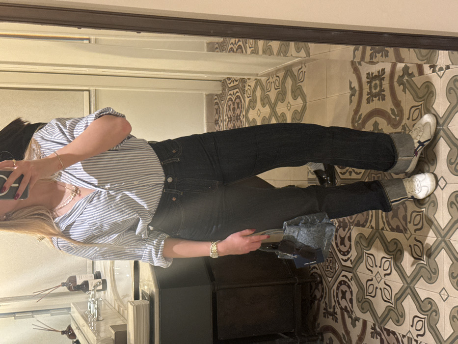 An individual is captured in a mirror selfie, presenting a full-body view from the upper chest down. The person has long blonde hair, a light blue and white striped button-down shirt with rolled-up sleeves, dark blue cuffed jeans, and white sneakers with beige and dark striped accents. A gold-colored watch is on the left wrist, a delicate gold-colored bracelet on the right, and a gold-colored necklace with a pendant is visible. Multiple rings adorn the fingers holding a black smartphone with a dark red case, which obscures the person's face. The left hand holds a small, dark denim-textured handbag with an attached tag and sunglasses.
The scene is an interior space, likely a bathroom or dressing area, characterized by a highly decorative floor composed of patterned encaustic tiles in shades of beige, grey, and dark brown, forming a complex geometric and floral motif. To the left, reflected in the mirror, is a dark grey or black vanity unit with a white sink, silver-colored cross-handle faucets, several brown glass bottles with reed diffusers, and a stack of white folded textiles. To the right, a plain light-colored wall and a doorway are reflected. The overall design aesthetic, particularly the distinct patterned flooring and fixtures, is consistent with renovated historic buildings, boutique hotels, or luxury residences found in areas such as Tel Aviv's White City or Neve Tzedek, Israel.