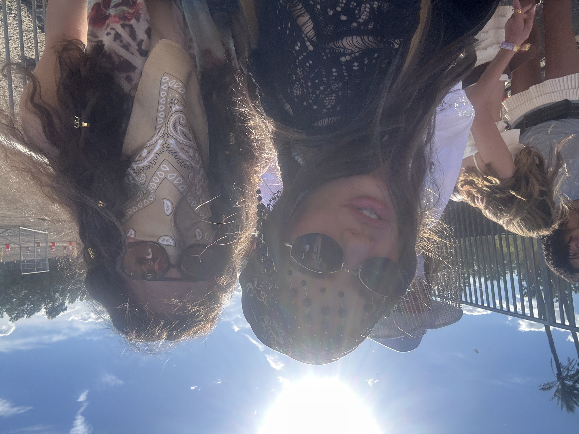 Two young women pose for a selfie on a sunny day at the Coachella Valley Music and Arts Festival in Indio, California, United States of America. The woman on the left wears sunglasses and a bandana. The woman on the right also wears sunglasses and a bandana with decorative beads. There are other attendees and trees in the background. The sun is bright and there is a blue sky with scattered clouds.
