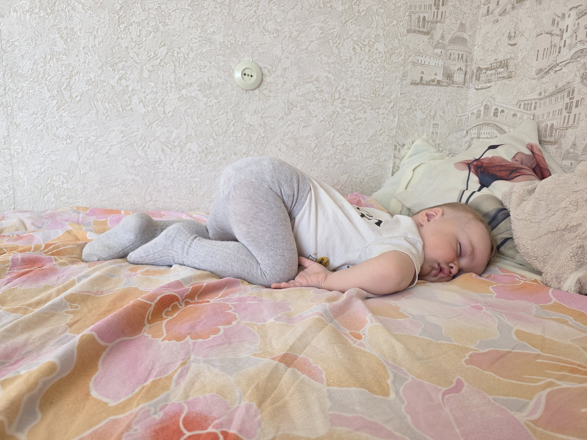 A baby is sleeping on a bed. The baby is wearing grey leggings and a white t-shirt. The bed has a floral-patterned bedspread in shades of pink, orange, and yellow. The baby is lying on their side with their face resting on the bed. A pillow with a floral design is behind the baby's head. The wall behind the bed has textured wallpaper and a scene of European buildings. An electrical outlet is visible on the wall. The location is Chaikovsky, Russia.