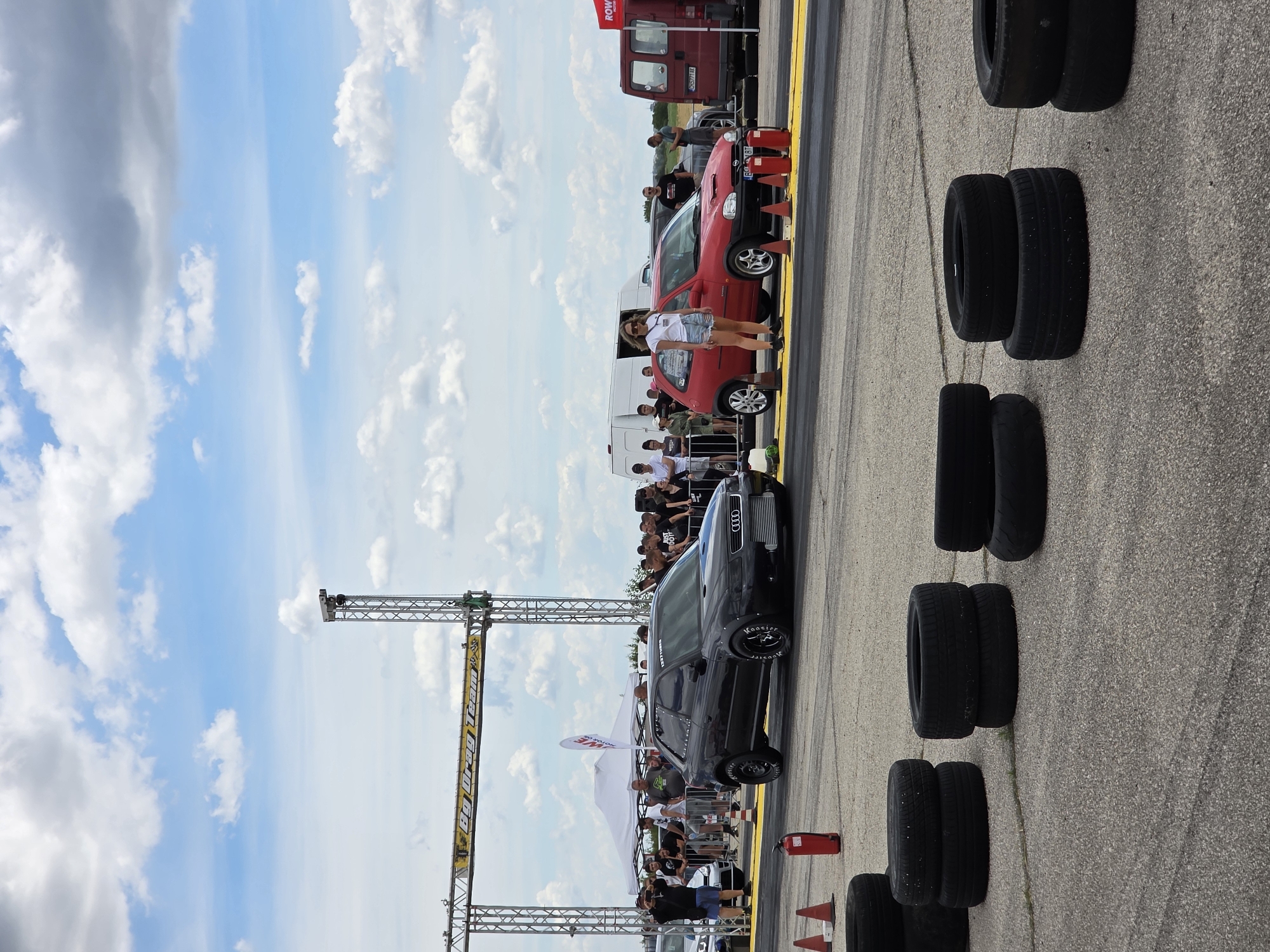 This image depicts a drag racing event at the Akatsievo airfield in Bulgaria. In the foreground, several tires are stacked on the asphalt. The middle ground features a drag racing track with a black Audi A3 and a red Opel Corsa positioned at what appears to be the starting line. Spectators are gathered behind a fence, watching the event. Several tents and vehicles, including a white van and a red truck, are visible in the background. The sky is partly cloudy.