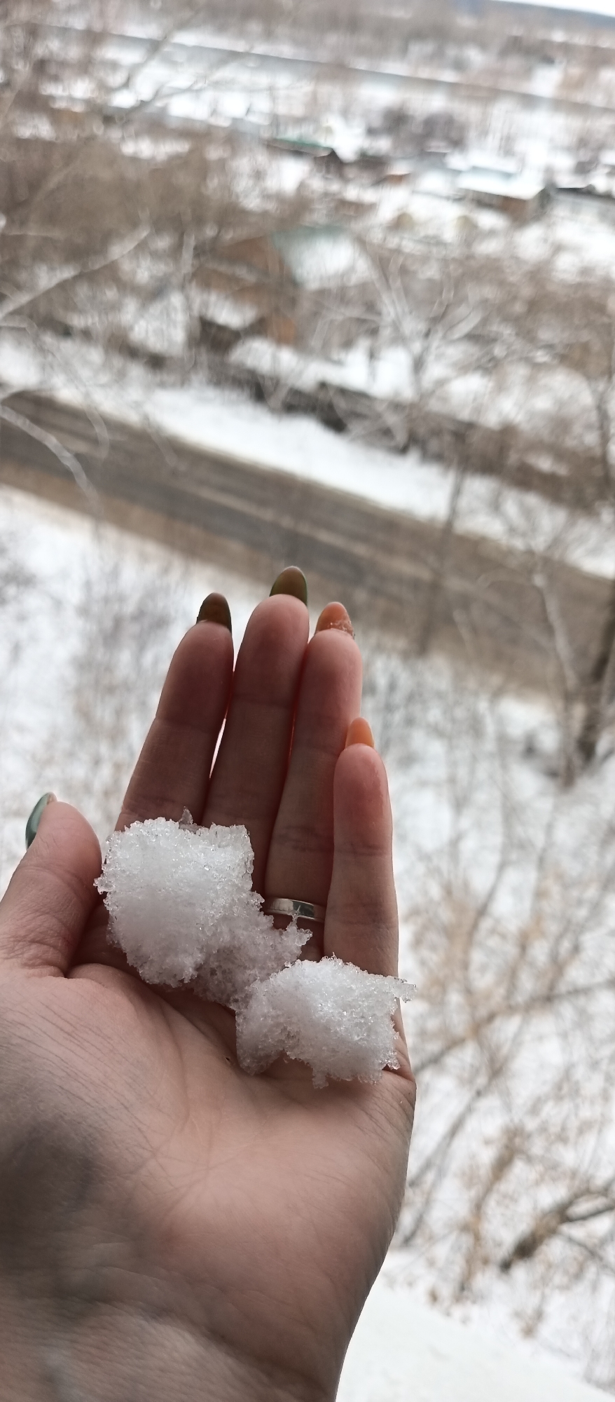 A high-angle medium shot depicts a hand holding snow against a blurred background of a city street on a snowy day. The hand, Caucasian, is palm-up, cradling a clump of snow. A silver ring is visible on the ring finger. The fingernails are painted with a gradient, ranging from green to tan to orange. The background features a street covered in slush, bare trees, and indistinct buildings covered with snow. The city and location are not identifiable from the available information.