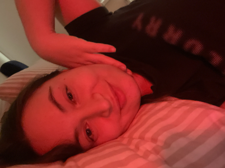 A young woman lies on a striped pillow, illuminated by red light. Her head is tilted, and she's gazing towards the camera with a gentle smile. Her hand rests near her head. She is wearing a dark t-shirt with light text printed on it. The background is simple and dark. The overall impression is intimate and relaxed.