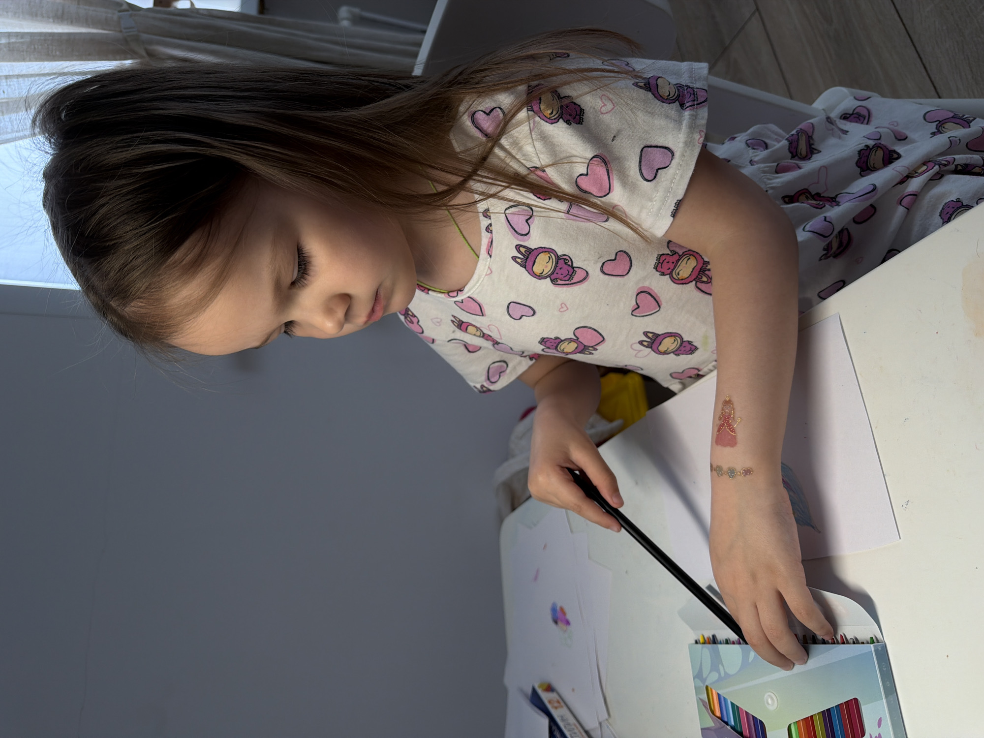 A young girl is seated at a white table. She is wearing a white shirt with pink hearts and cartoon characters. She is drawing with colored pencils. On the table, there are several sheets of paper with drawings and a box of colored pencils. The location is Chaikovsky, Russia.
