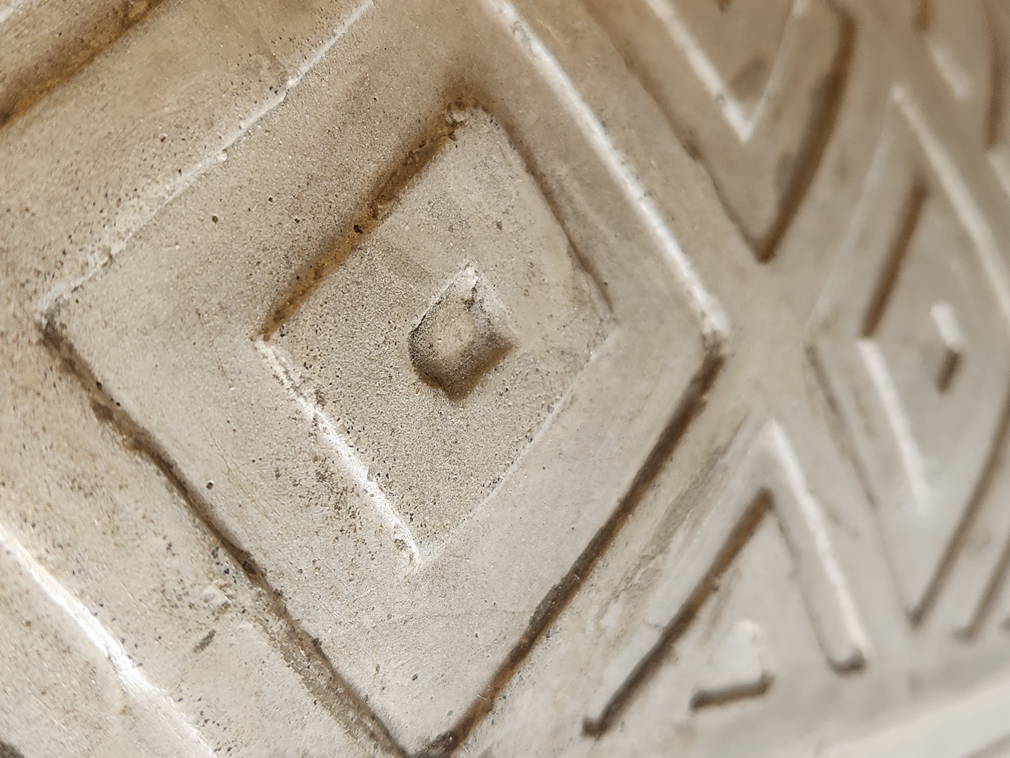 This is a close-up, low-angle, macro photograph of a rough, textured, concrete surface with a repeating diamond pattern. The concrete is a light beige or off-white color and shows signs of wear and some dark staining or dirt. The pattern consists of concentric squares that form diamonds. The lighting creates shadows that highlight the texture and relief of the pattern. The image appears to be an abstract detail of an architectural element or perhaps the sole of a shoe or a decorative object. There are no people, actions, or specific environmental context visible in this abstract close-up. Without additional information, it is impossible to determine the location in Maitland, United States of America.