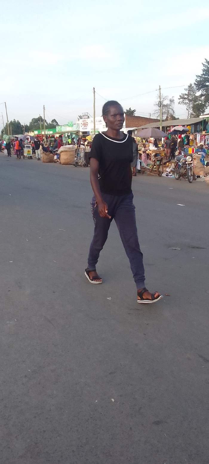 A dark-skinned woman with short, braided hair is walking across a paved road from left to right. She is wearing a black short-sleeved t-shirt with a white trim around the collar, dark blue sweatpants, and dark brown sandals with white soles. Her expression is neutral as she looks forward.
The scene behind her reveals a busy outdoor market, typical of Kakunga, Kenya. Numerous market stalls are set up along both sides of the road, displaying various goods. Many people are visible in the background, engaged in selling, buying, or moving through the market. Some individuals are attending to their stalls, while others are pushing handcarts or standing near parked motorcycles. Large sacks of goods are placed near the stalls, indicating a commercial area. Utility poles line the street, and buildings are visible beyond the market structures.
The lighting suggests either early morning or late afternoon, with a bright, clear, and slightly hazy sky, indicating fair weather. On a building in the mid-background, part of a sign is visible with the text "CYBER" and "HOURS", and another partially obscured sign appears to read "The Lou...". The overall atmosphere is one of a lively and active community market.