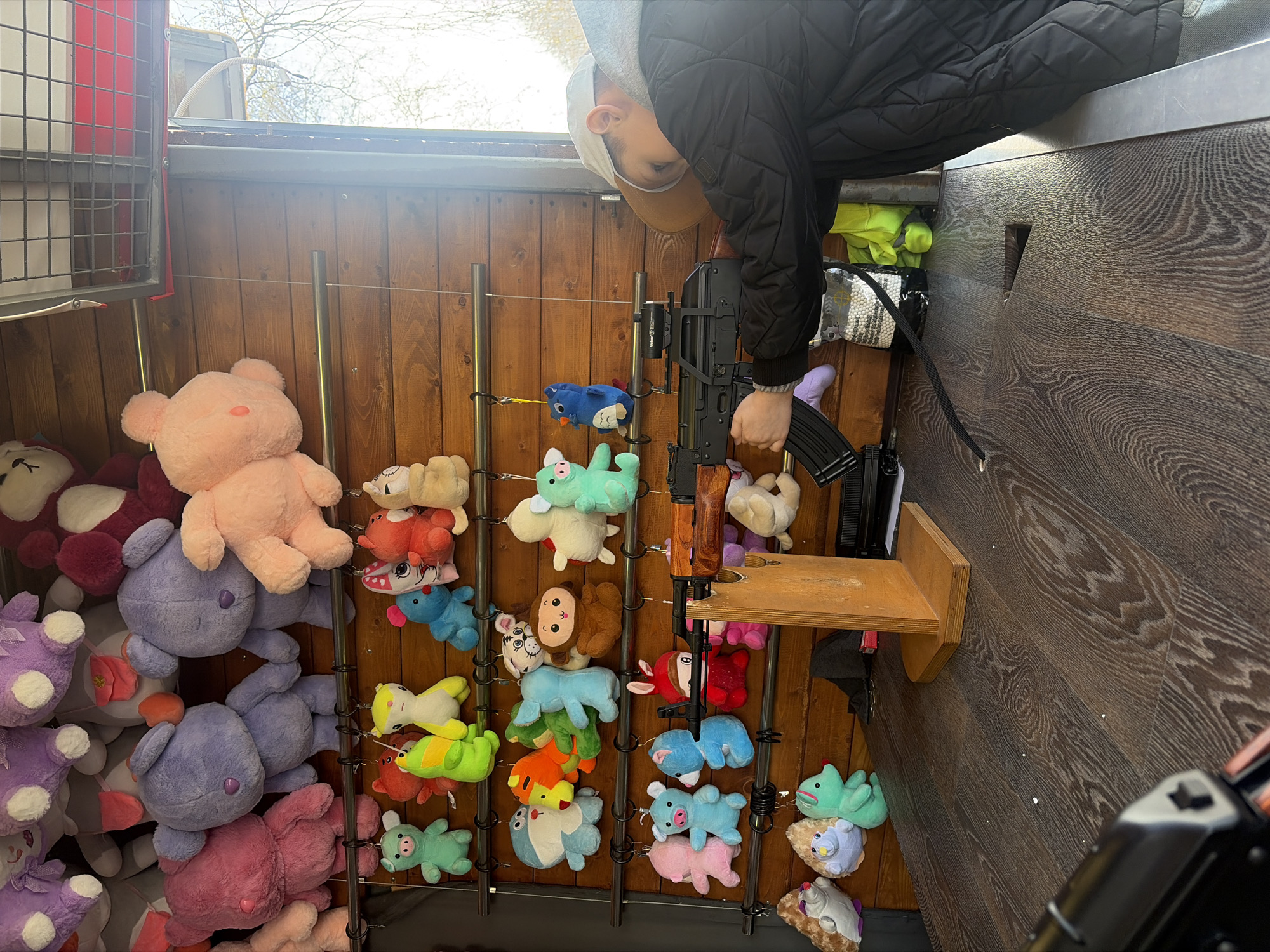 The image depicts a scene inside a shooting gallery booth, likely at a carnival or fair in Saint Petersburg, Russia. A young person, possibly a child or teenager, dressed in a dark quilted jacket, a grey hoodie, and a light-colored baseball cap, is captured leaning over from above. They are actively engaged in playing, holding a replica rifle, which appears to be an AK-style airsoft or pellet gun, equipped with a scope or laser attachment. The rifle rests on a small, light-colored wooden shelf attached to the booth's structure.
The background of the booth features vertical wooden paneling, against which numerous plush toys are displayed as targets. These stuffed animals come in various sizes and colors, including large pink and purple teddy bears, as well as smaller figures like pigs, frogs, monkeys, and elephants, all hanging from hooks on metal rods. On the floor of the booth, bags filled with white pellets, likely ammunition for the rifle, are visible.
Above the booth, a glimpse of the outside environment shows bare tree branches against a bright sky, indicating it is daytime. A metal grate or cage structure is also visible on the upper left side of the booth. The scene captures the participant's focused attention on the game, aiming the replica firearm at the array of plush prizes. There is no visible text within the image.