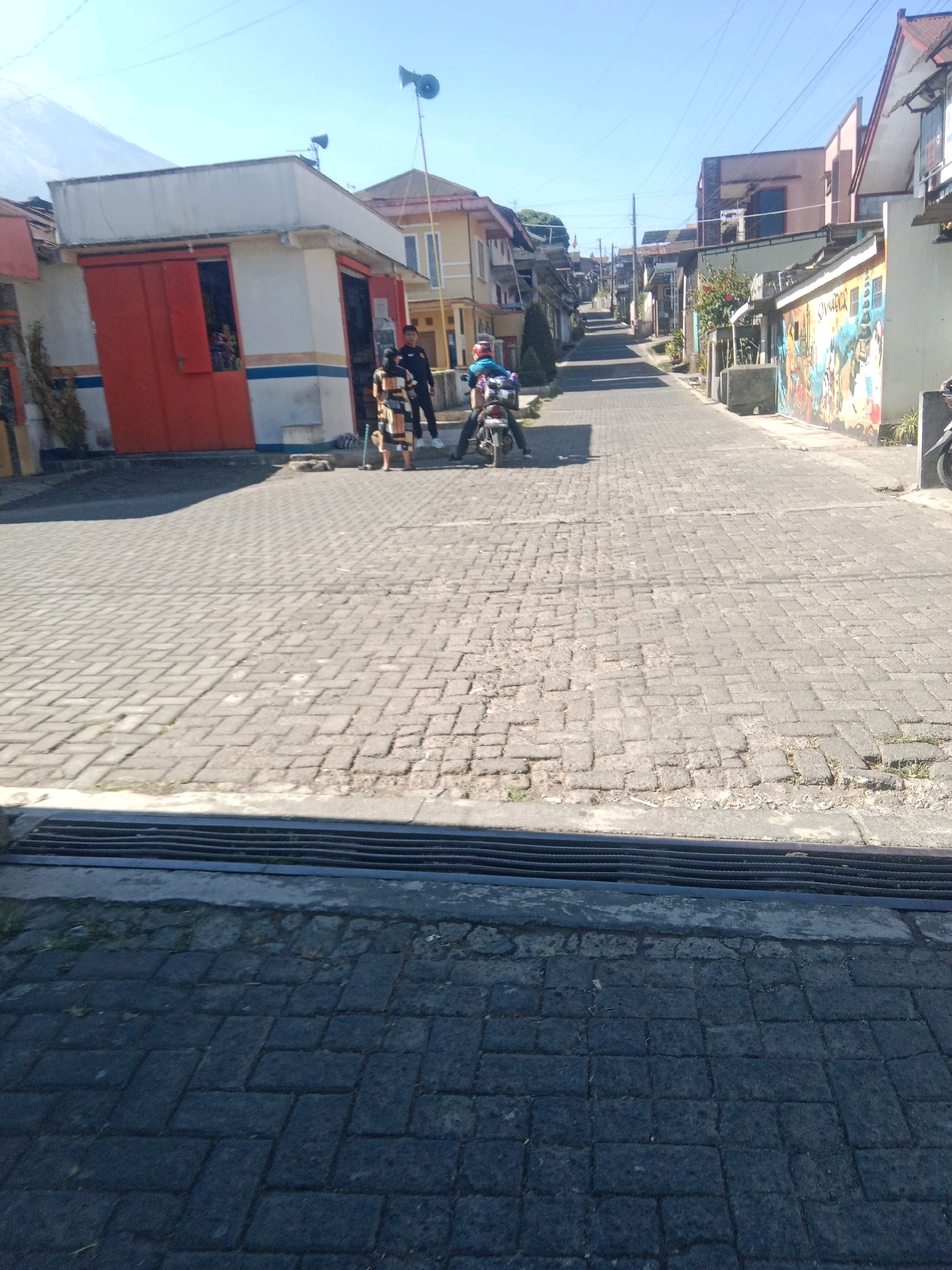 The image depicts a street scene in the city of Kapencar, Indonesia, on a clear, sunny day. The street is paved with cobblestones. In the foreground, a drain grate runs horizontally across the bottom of the frame. To the left of the street, a white building with red doors and blue and orange trim is visible. Two individuals, one wearing a patterned dress and the other in a dark uniform, stand near the building. To the right of them, a person on a motorcycle wearing a blue helmet is also present. The street extends into the distance, lined with buildings and houses on both sides, some of which feature colorful murals. Overhead power lines are visible against the bright blue sky.