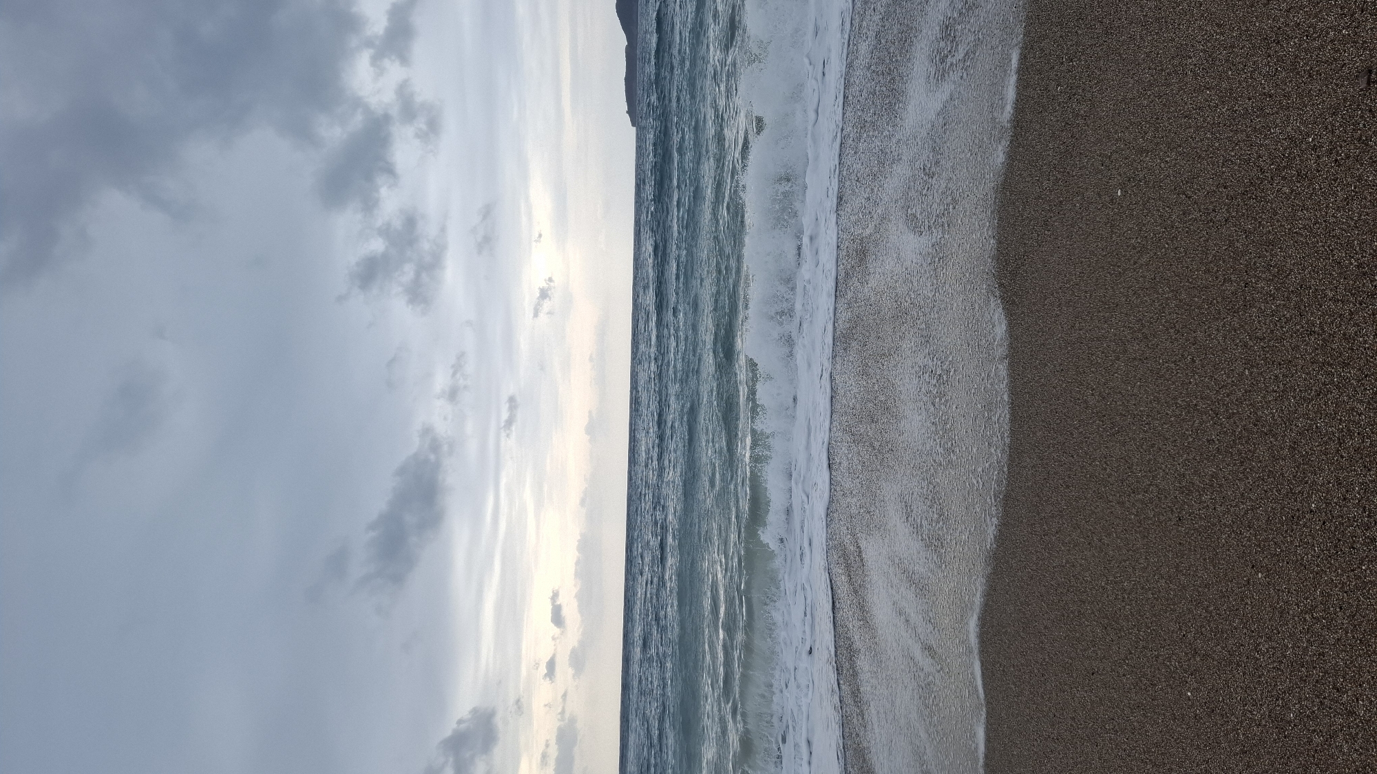 The image presents a wide view of the coastline in Boreti, Montenegro, on an overcast day. The foreground displays a dark, wet pebbly beach, occupying approximately the bottom third of the frame. Immediately beyond the beach, moderate waves are visible breaking with white foam as they reach the shore. The sea exhibits deep blue and greenish-turquoise tones. The horizon line bisects the upper two-thirds of the image, separating the water from the sky. The sky is largely covered by varying shades of grey clouds, with a brighter, lighter grey area visible near the horizon in the center. A dark, elevated landmass or promontory is discernible on the far right, extending into the sea. No individuals or man-made objects are present.
