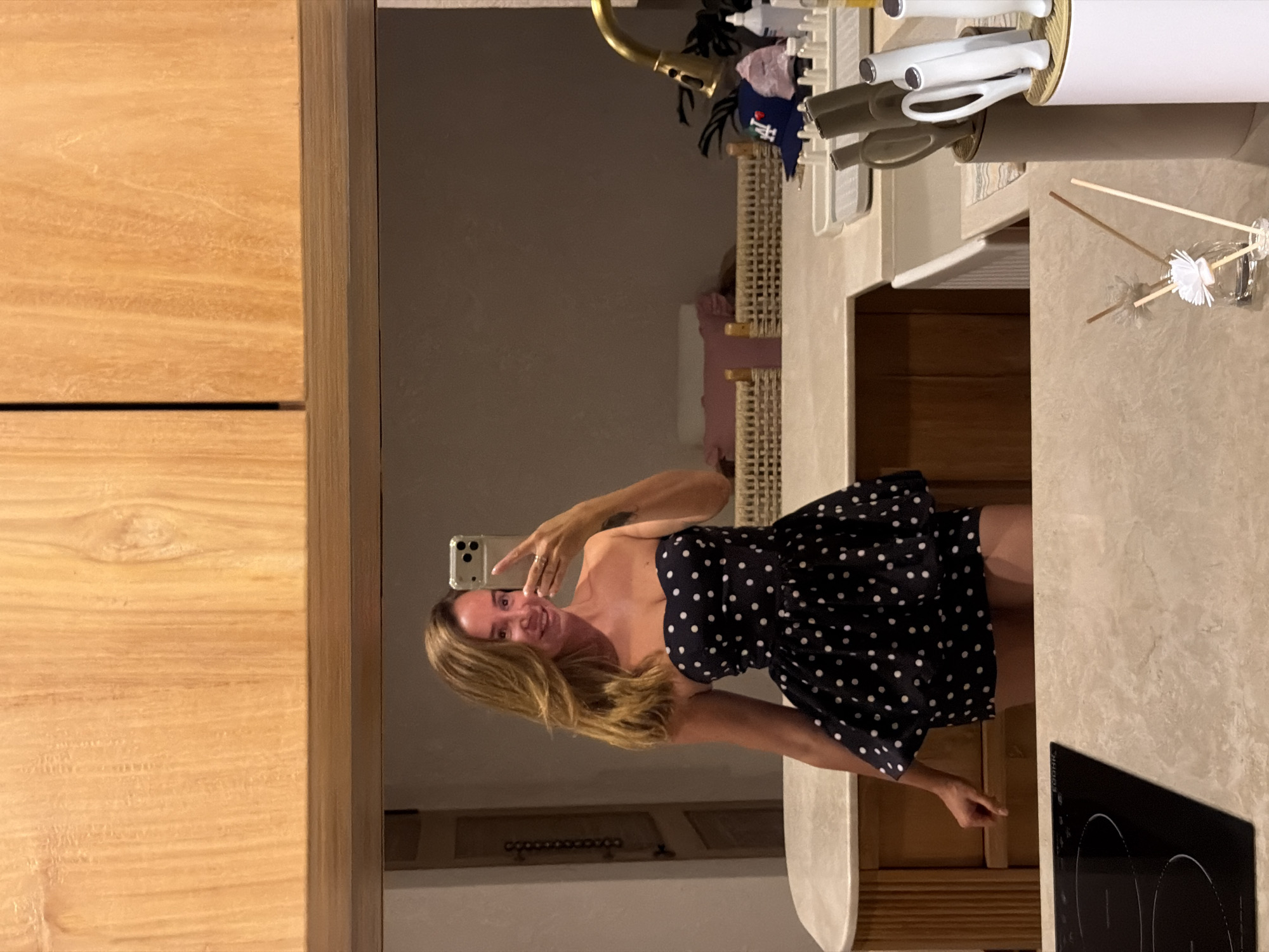 A woman is reflected in a mirror, taking a selfie. She is wearing a strapless, dark blue dress with white polka dots, and she is smiling at the camera. Her blonde hair is styled down, and she is wearing rings on her fingers. The background appears to be a modern kitchen with wooden cabinets, a countertop, and a sink. There are various kitchen items visible, including utensils in holders, a faucet, and a bottle of cleaning product. A framed picture or artwork is also visible in the background. The lighting suggests an indoor setting, possibly a home or a restaurant.