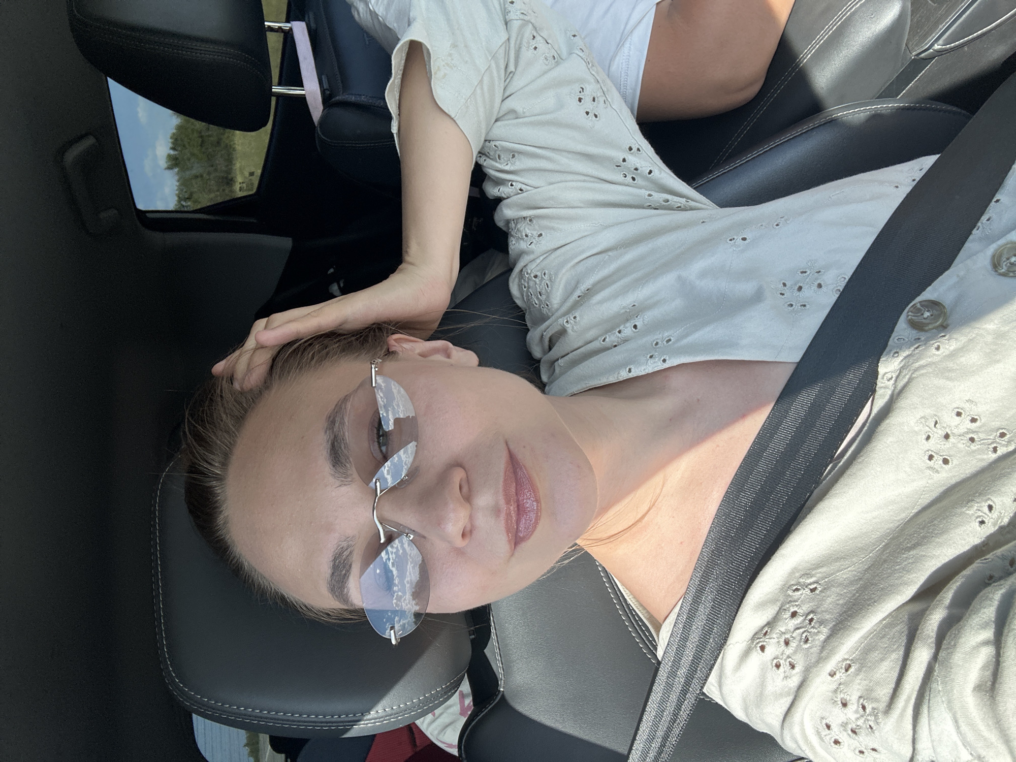 The image shows a woman wearing sunglasses and a light-colored, embroidered shirt, sitting in the passenger seat of a car. She is resting her hand on her head, and her arm is extended behind her. The car appears to be in motion, as indicated by the reflection of clouds on her sunglasses and the scenery visible through the rear window. The background suggests a rural or suburban environment, with trees and a blue sky. There is no discernible signage or landmark to indicate the specific location within Antipovka, Russia. The seatbelt is fastened across her chest. Another person is partially visible in the background, to the right of the woman.