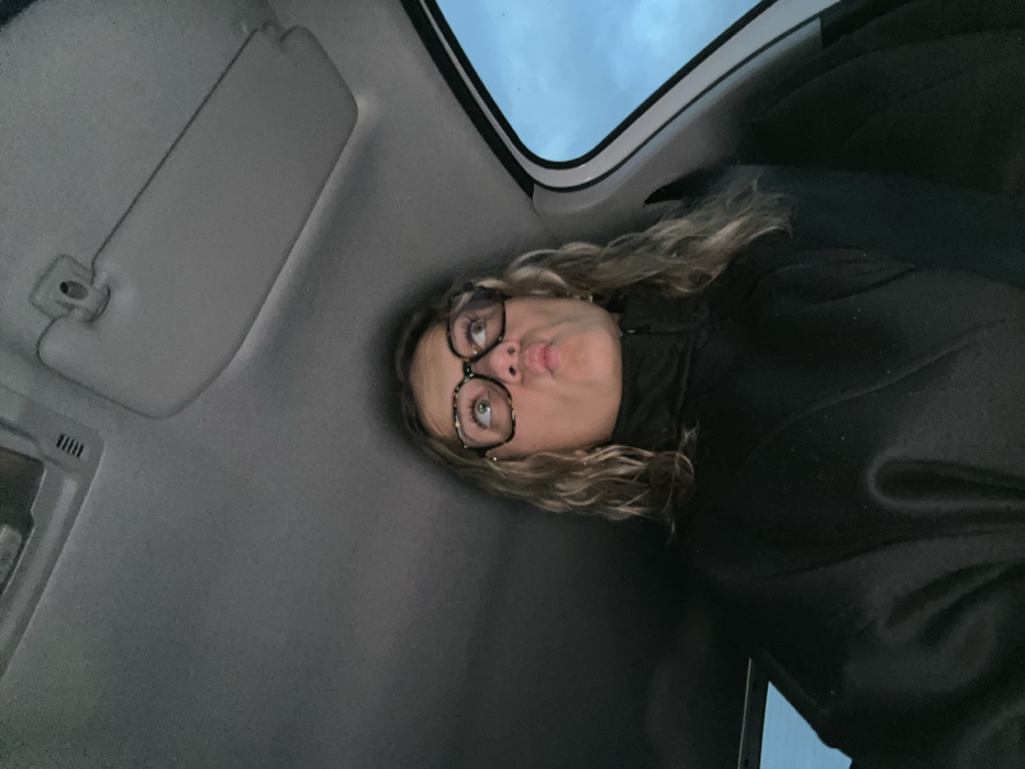 A person wearing glasses and a black jacket looks upward, puckering their lips, inside of a vehicle. The car's interior includes a gray ceiling, sun visor, and a cloudy sky visible through the window. The image appears to be taken at an angle, with the person positioned lower than the camera.