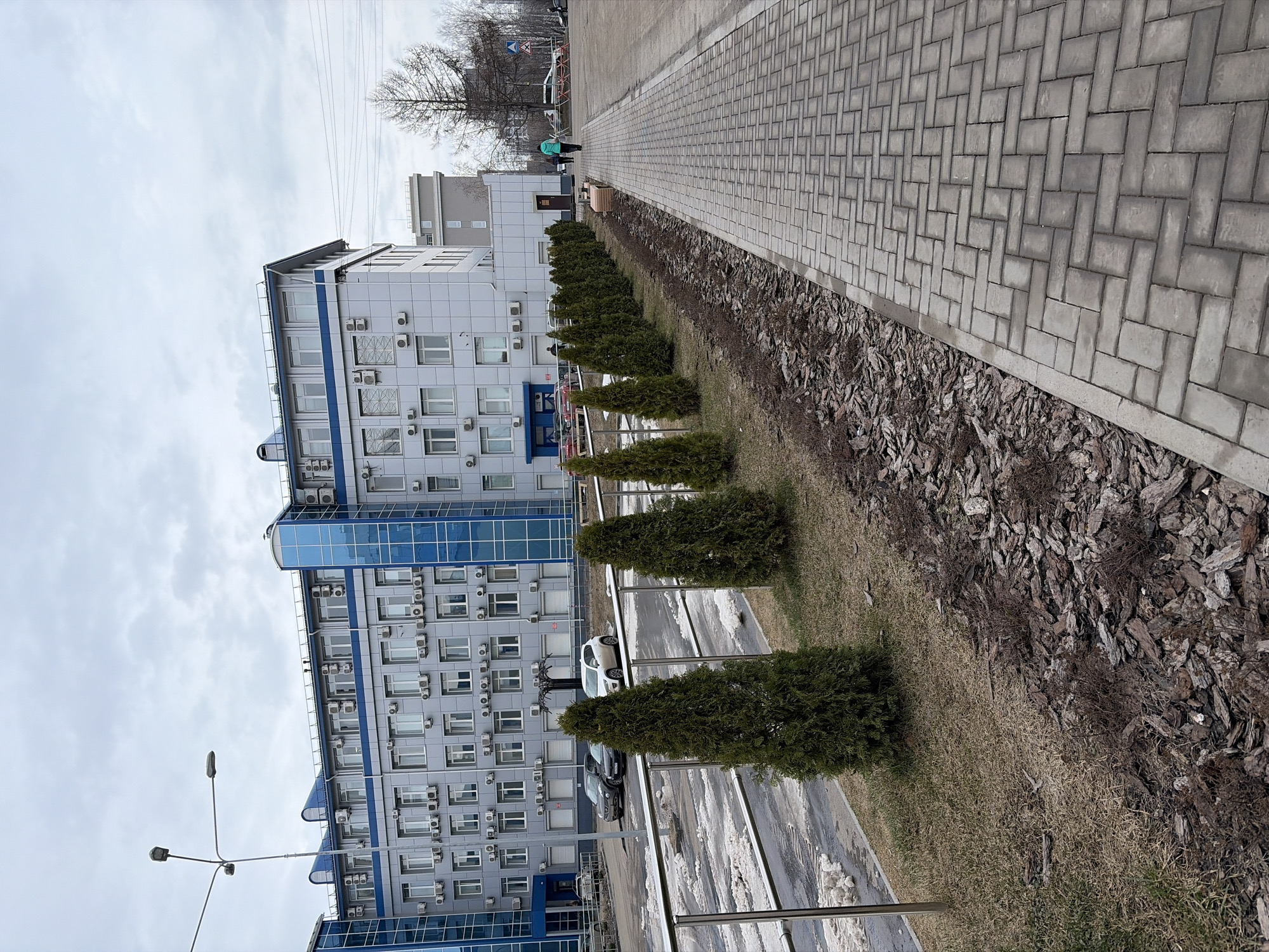 The image depicts an exterior view in what appears to be an urban setting. A multi-story building with a blue and gray facade is present, featuring numerous windows, each equipped with what seems to be individual air conditioning units. A row of manicured, cone-shaped evergreen trees lines the edge of a wide walkway or sidewalk paved with interlocking gray bricks. A strip of ground cover, composed of mulch and dry grass, separates the trees from the paved area.
Several cars are parked along the side of the building. Streetlights are visible in the foreground. The sky is overcast, suggesting either early morning or a cloudy day.
Based on visual cues and provided context, the location is identified as Izhevsk, Russia. There are a few people walking along the sidewalk in the distance.