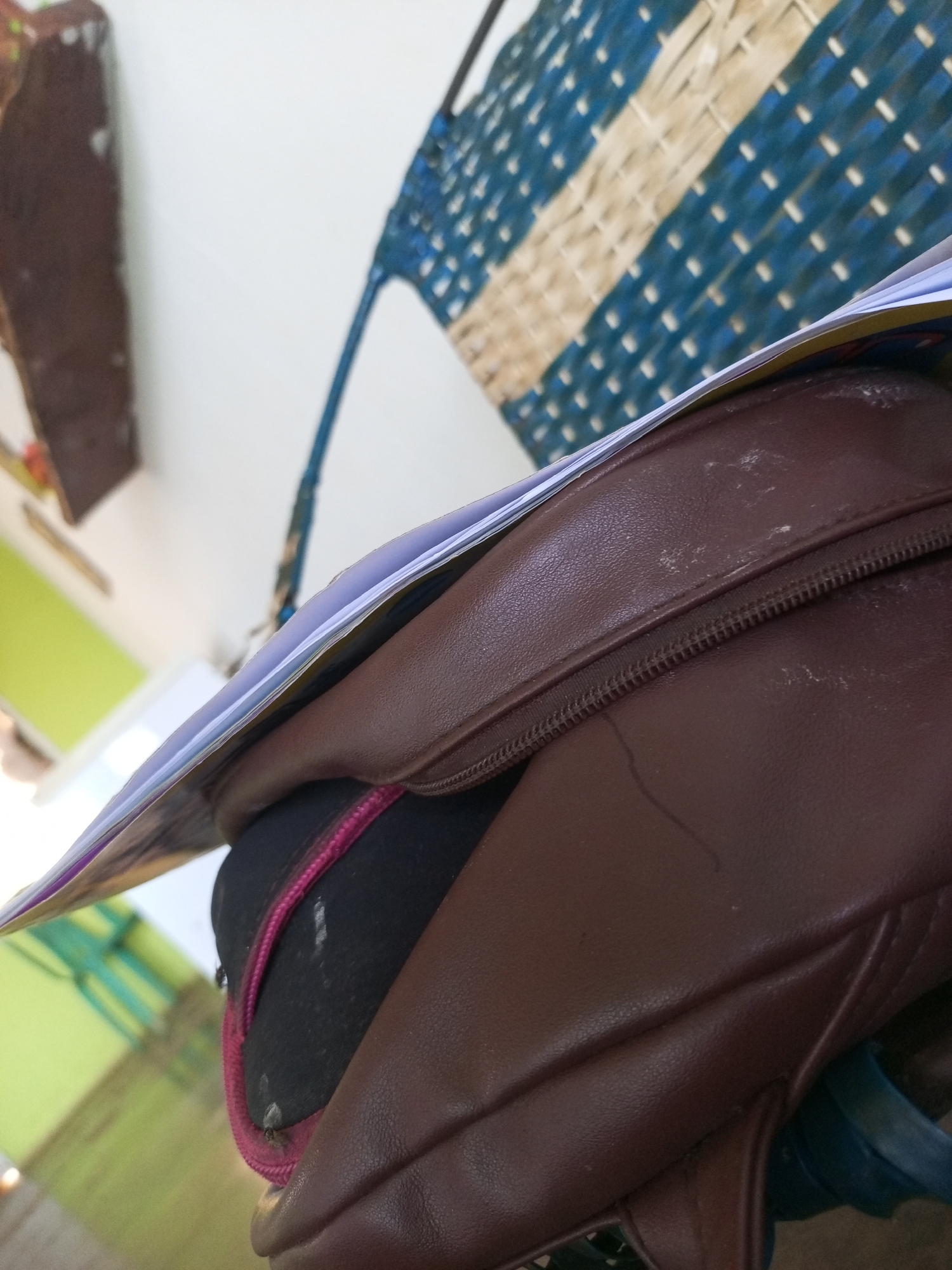 A brown leather bag with a pink zipper is resting on a surface, with a stack of papers on top of it. In the background, a woven blue and white mat hangs on a white wall. The scene appears to be indoors, possibly in a home or school in La Tagua, Venezuela. The lighting is natural, coming from an unseen window, casting soft shadows on the objects. There are no people visible in the image. The overall atmosphere is calm and ordinary, suggesting a moment of everyday life captured in a candid shot.