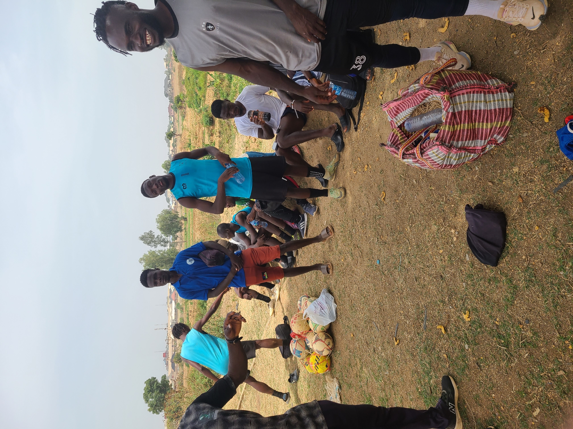 A group of young men are gathered outdoors in Barikin Jirga Summa, Nigeria. They appear to be resting, possibly after a soccer game. Several soccer balls are piled together. Some of the men are holding water bottles. One man is sitting with a dark-colored jar in hand. A colorful, striped bag lies on the ground. The ground is a mix of dry dirt and sparse grass. Buildings can be seen in the background.