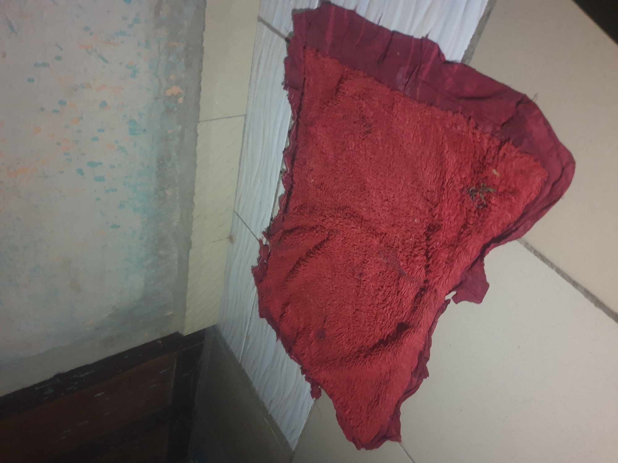A red, frayed, and somewhat dirty rag lies on a tiled floor in what appears to be an indoor setting, possibly a hallway or a room. The rag is centrally located in the frame, angled diagonally. The floor is covered in light-colored square tiles with visible grout lines. To the left of the rag, a dark wooden door or frame is partially visible. The wall in the background is textured and painted with light colors, exhibiting some splotches of light blue and orange. The scene is dimly lit. There are no people or animals visible in the image.