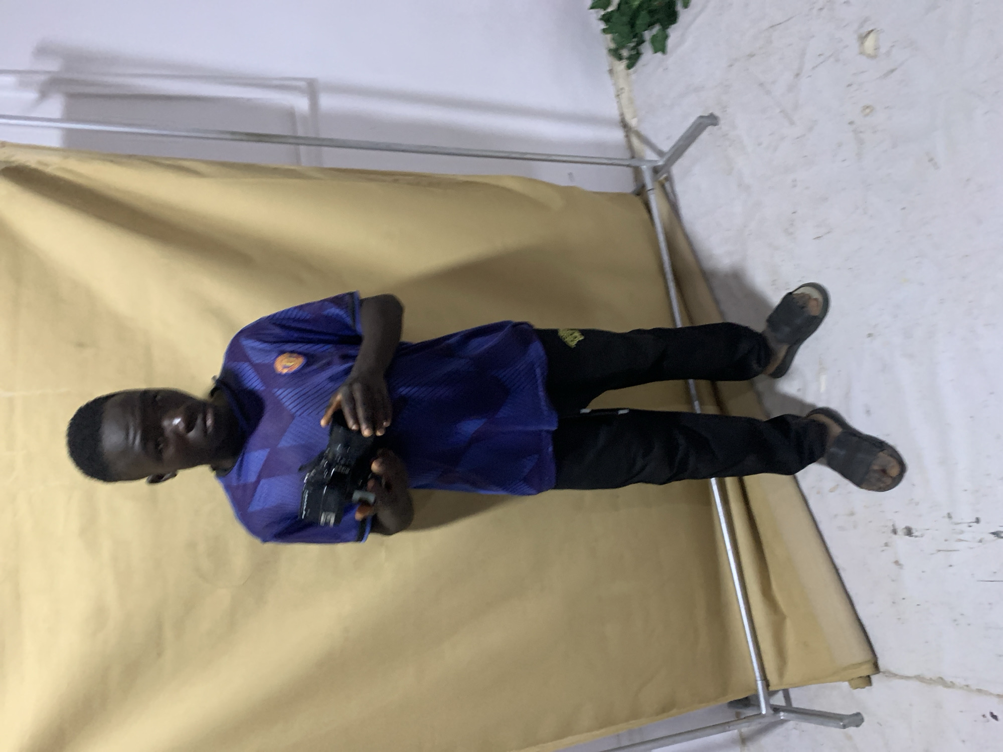 A Black man stands holding a camera. He wears a blue and purple shirt and black pants. He also wears black sandals. He stands in front of a beige backdrop that is held up by a metal frame. The background appears to be a wall. Greenery can be seen above the backdrop. There is no evidence to confirm or deny that the location is in the city of Jos, Nigeria.