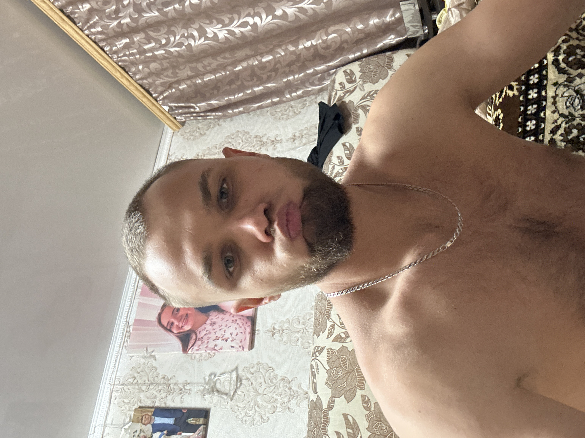 A young, shirtless man with short hair and a beard is taking a selfie in a room. He is puckering his lips and looking directly at the camera. He is wearing a silver chain necklace. In the background, there is a patterned wallpaper, a sofa with a floral pattern, and a large window with draped curtains. Two framed pictures are hanging on the wall above the sofa. The overall impression is that of a casual, personal moment captured in a domestic setting. There is no indication of the location being in Загородный, Russia.