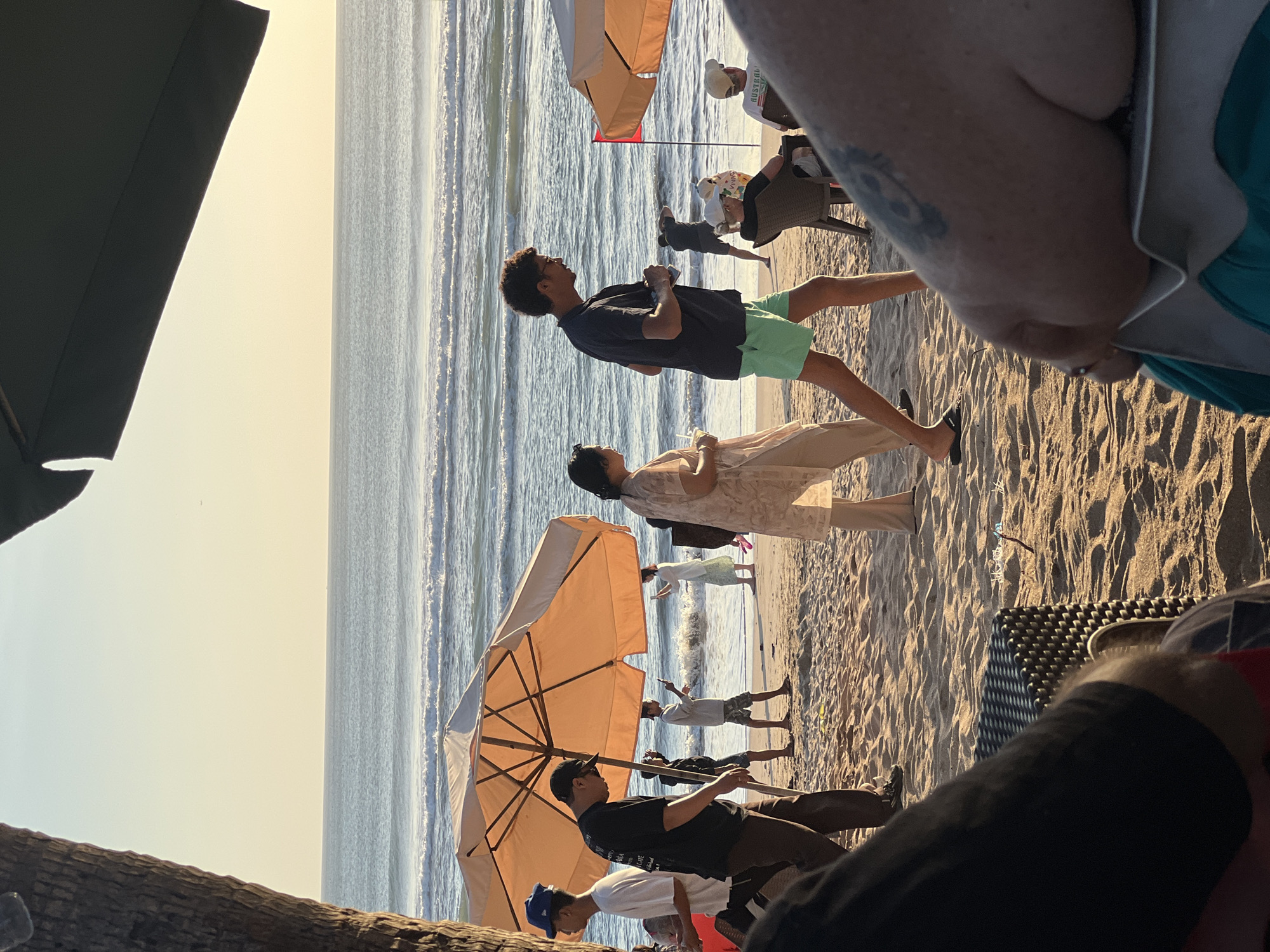 This is a photograph of Kuta Beach in Kuta, Indonesia, at sunset. The sky is a pale yellow, and the ocean has gentle waves. Several people are on the beach, some walking, some sitting.
In the foreground, two people are seated on chairs, their backs to the camera. One person has a tattoo on their arm. A large beach umbrella shades a portion of the sand.
Further back, several individuals are visible. A man in a dark t-shirt and light green shorts walks towards the right side of the frame, his profile visible. To his left, a woman in a light-colored, loose-fitting outfit is also walking. Other people are scattered across the beach, some near the water's edge. One person appears to be taking a photograph of the ocean with their phone. A red flag is planted in the sand near the water. The sand is light brown and appears dry, with some scattered debris. The overall atmosphere is relaxed and casual.
