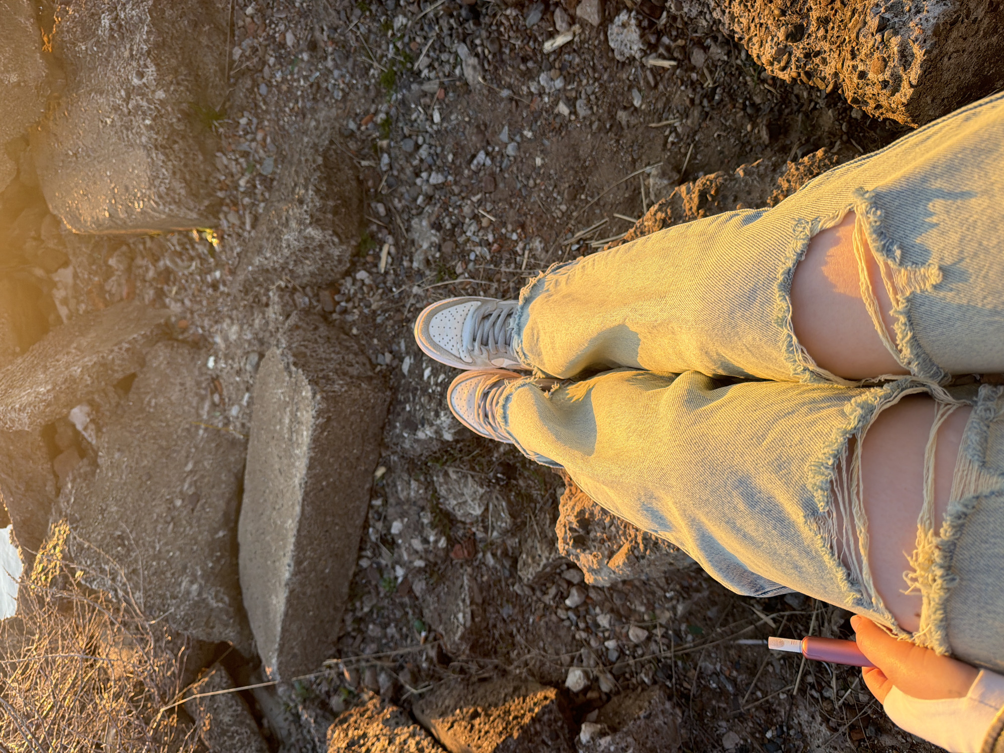 The image, taken from an elevated, first-person perspective, shows a person standing outdoors in Chaikovsky, Russia. The person is wearing ripped light blue jeans and white sneakers with pink soles. The surface underfoot consists of broken concrete, small stones, and dry vegetation. In the bottom right corner, the person is holding a pink electronic device. The scene is lit by golden hour sunlight.