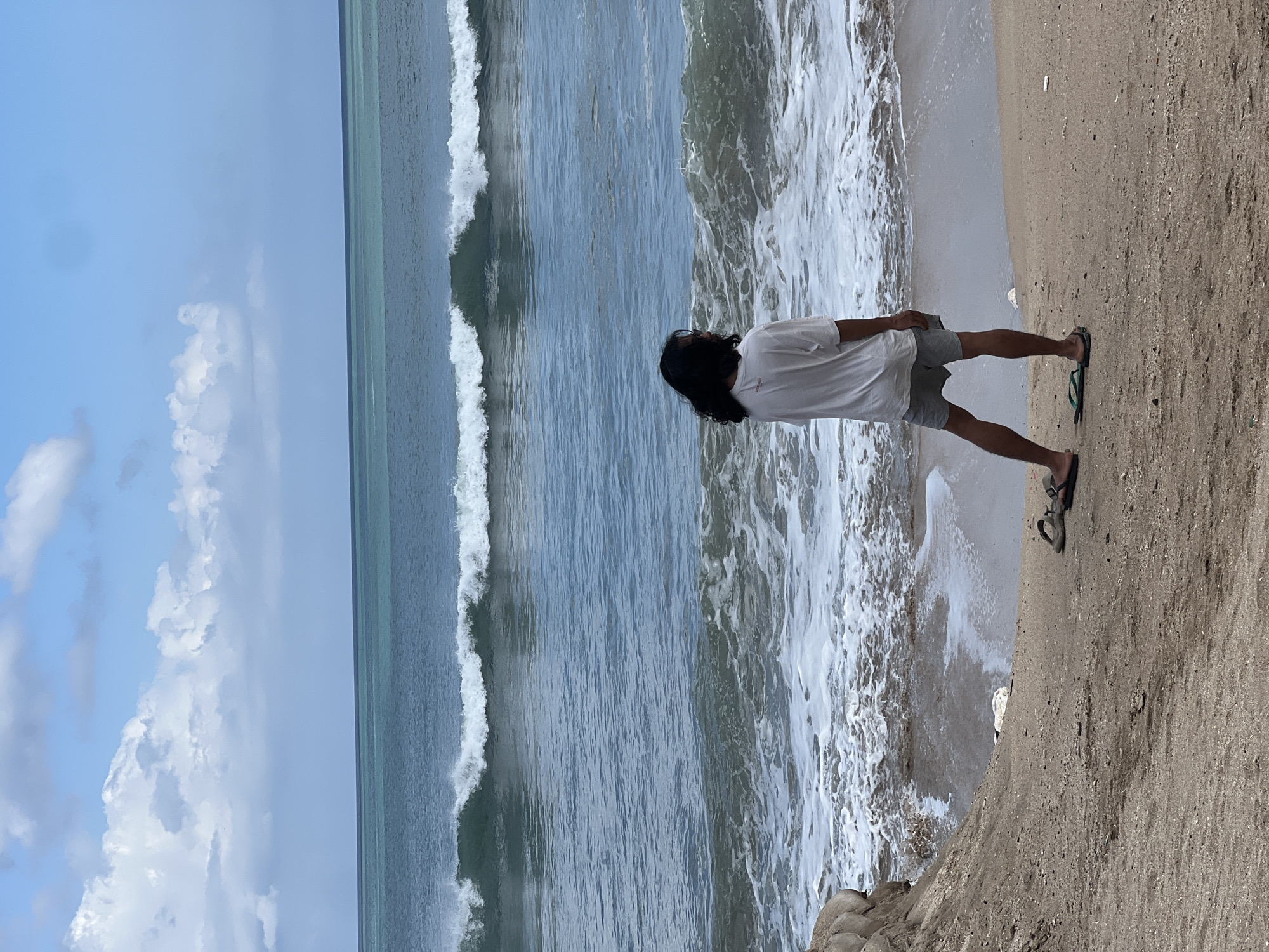 A person with long, dark hair stands on a sandy beach, facing the ocean. They are wearing a white t-shirt and gray shorts. Flip-flops are on the sand near their feet. The ocean has waves breaking on the shore, and the sky is blue with white clouds. The location is Kuta, Indonesia.