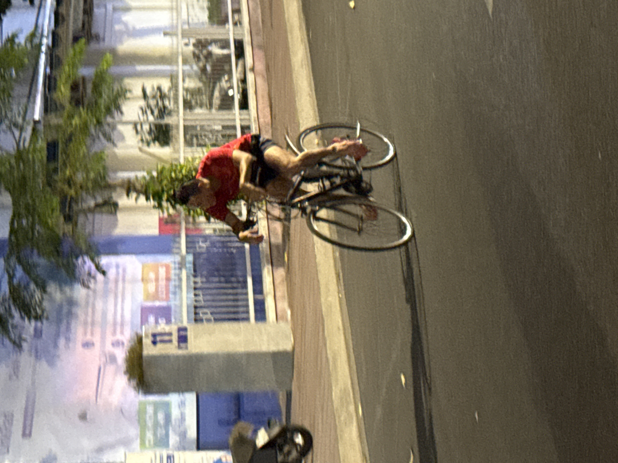 A person in a red shirt and dark shorts is performing a wheelie on a bicycle. The scene appears to be a street in Nha Trang Ward, Vietnam, as indicated by the text and the general atmosphere. The person's body is angled, with their legs extended behind them to maintain balance. The bicycle is positioned upright, with both wheels in motion, suggesting a dynamic and skilled maneuver.
The background shows a building with large windows and some greenery. The lighting suggests it's either early morning or late evening, likely during twilight hours, as there are some streetlights or ambient lights illuminating the scene. The pavement is visible, and a darker road is to the right. There are various posters or signs on the building in the background. One sign has text that includes the word "Vietnam" and what appears to be Vietnamese characters. Another sign is blue and white and has the number "11" on it. The overall impression is an urban environment. The activity suggests an individual practicing or showcasing a cycling skill. There are no overt emotional cues visible, but the act of performing a wheelie typically implies confidence and focus.