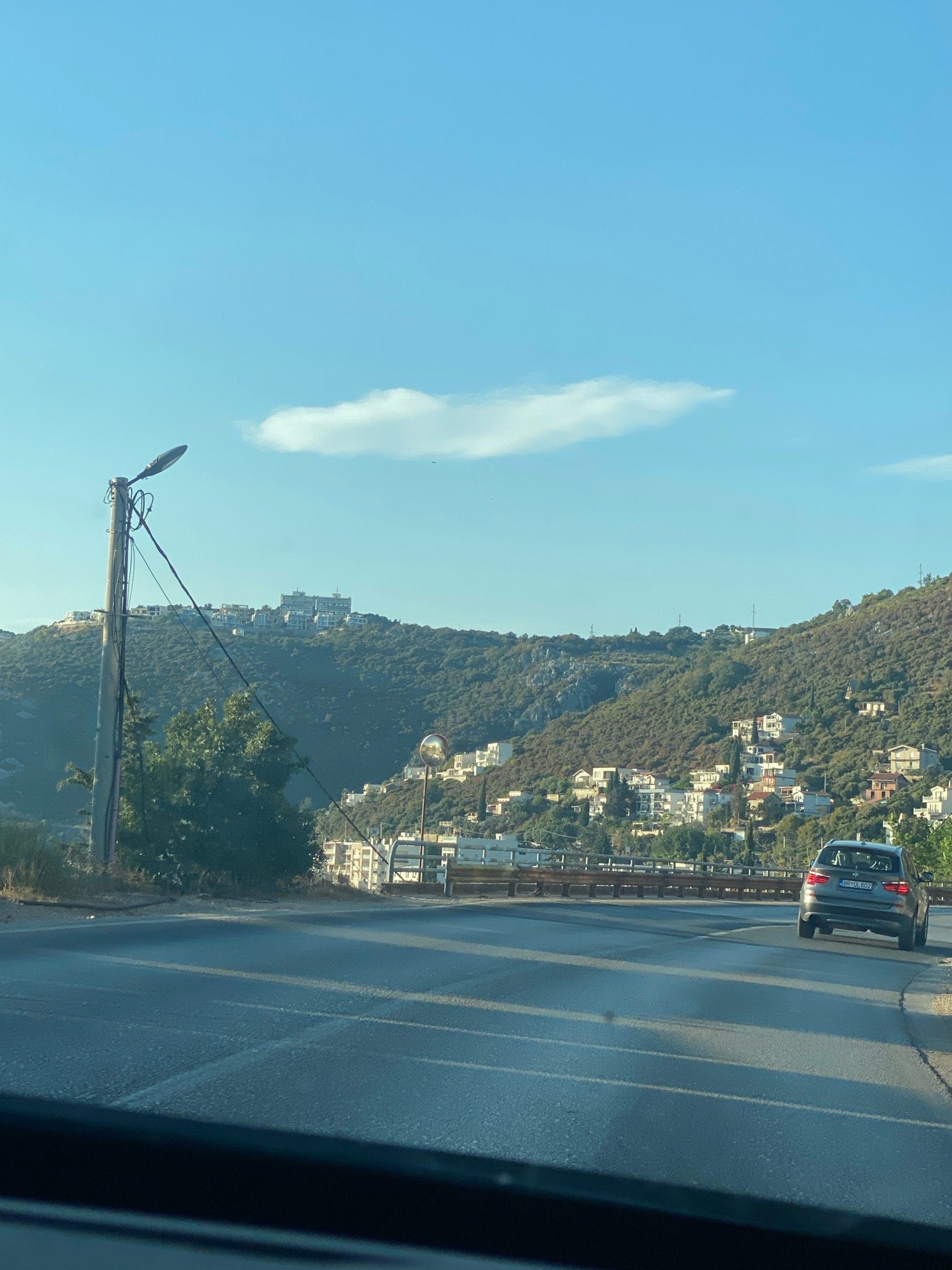 The image depicts a view from a vehicle driving on a road in Dobra Voda, Montenegro. In the foreground, the asphalt road is visible, with tire tracks and lane markings. On the right side of the road, a gray SUV is driving away from the camera, displaying a license plate that reads "BR DL 802". Beyond the road, a hillside is covered with green vegetation and dotted with white buildings. A bridge railing runs parallel to the road. On the left side of the road, a tall lamppost with electrical wires is prominent. In the background, more buildings are scattered across the hills under a clear blue sky with a single, elongated cloud. The overall scene suggests a sunny day in a coastal town.