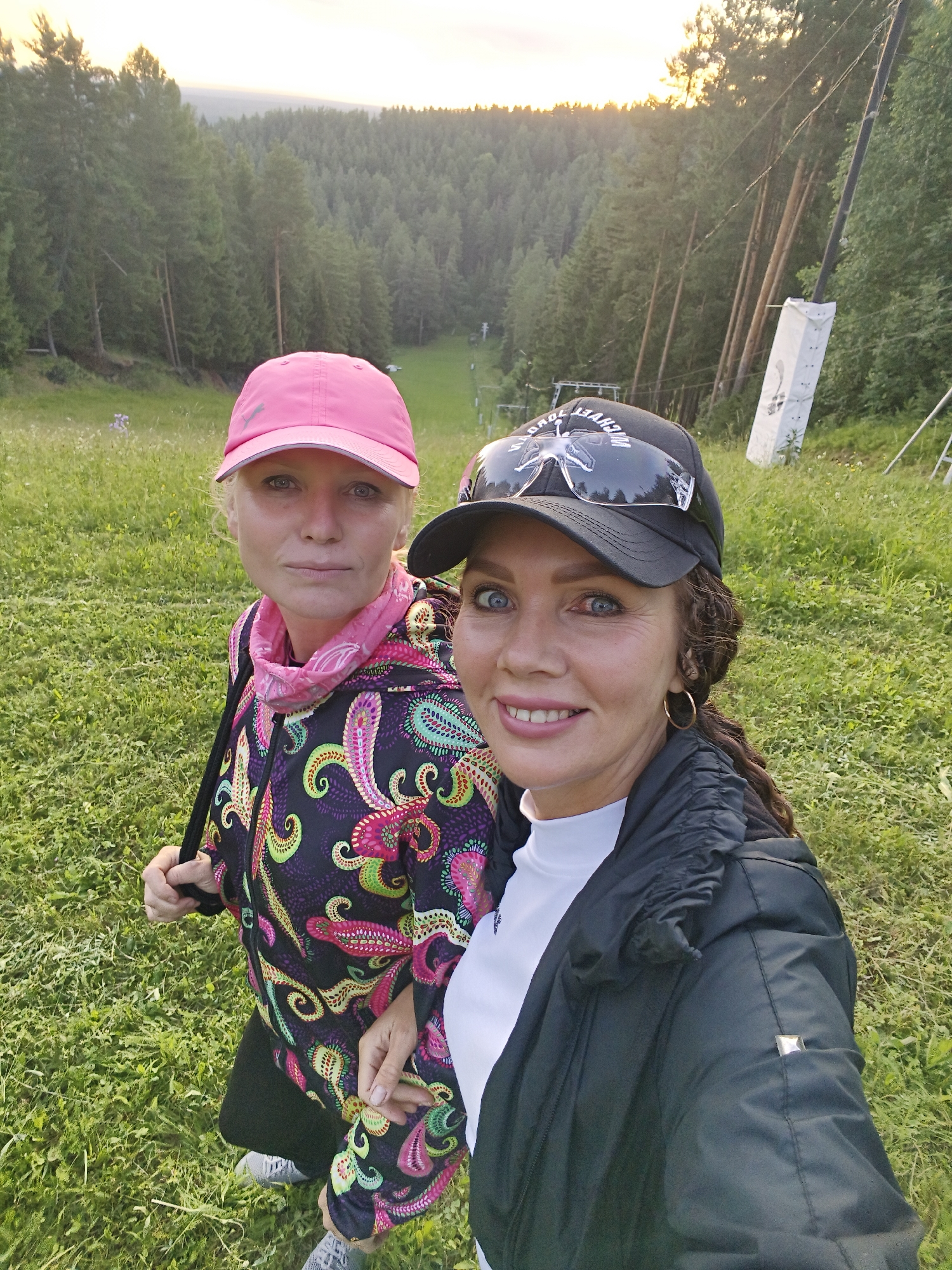 Two women are posing for a selfie in a grassy, wooded area. The woman on the left is wearing a pink baseball cap, a colorful paisley print zip-up jacket, and a pink bandana around her neck. She has blonde hair and is looking at the camera with a neutral expression. The woman on the right is wearing a black baseball cap with sunglasses perched on top, a black puffer jacket over a white turtleneck shirt, and gold hoop earrings. She has dark curly hair and is smiling broadly at the camera. The background consists of a dense forest of tall evergreen trees, with a ski slope or clearing visible in the distance. The sky is bright and hazy, suggesting it is either sunrise or sunset. The overall impression is of an outdoor recreational setting, possibly a mountain resort.