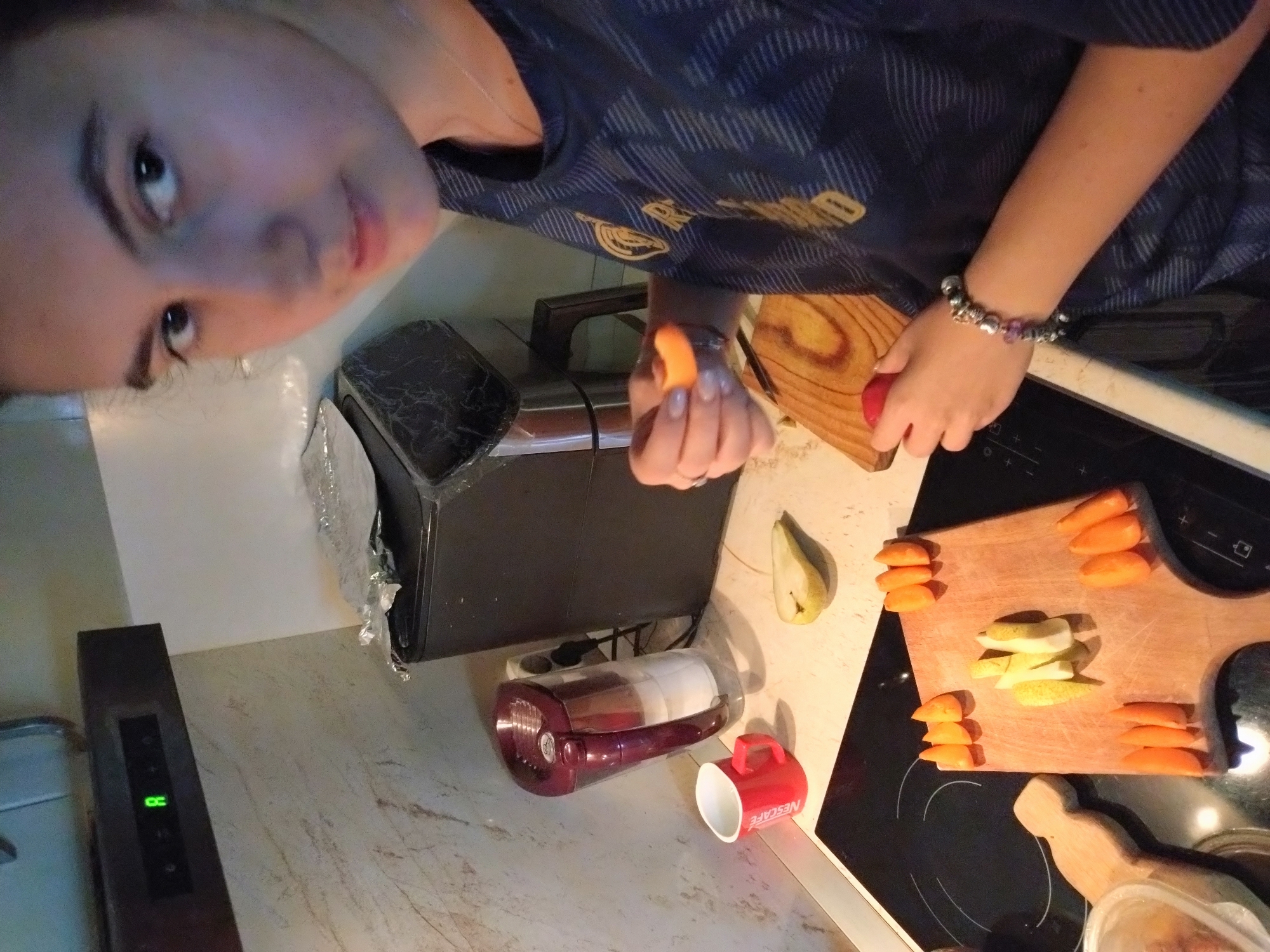 A young woman is in a kitchen, likely in Stara Zagora, Bulgaria, preparing food. She is standing in front of a black electric stovetop, and to her left is a red mug and a water filter pitcher. An air fryer is visible in the background, and above it, a range hood with a green digital display showing the letter 'R'. The woman is holding a slice of orange fruit in her right hand and is looking towards the camera. In front of her, on a wooden cutting board, are sliced carrots and pears. She is wearing a dark blue t-shirt with a pattern and a bracelet on her left wrist. The overall environment appears to be a domestic kitchen setting.