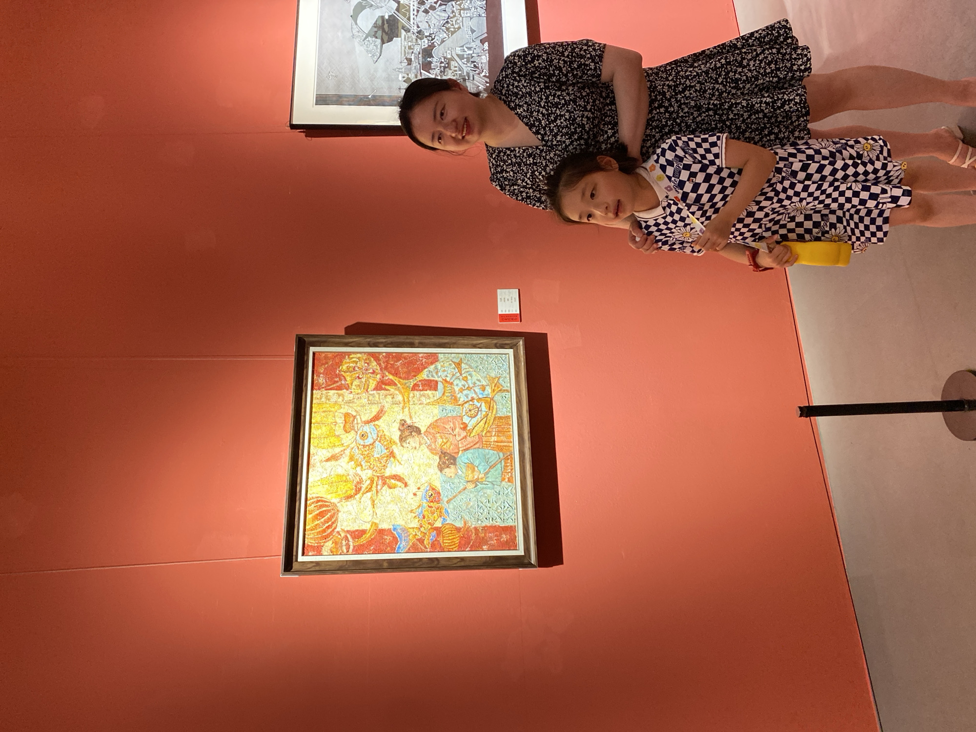 A woman and a young girl stand in front of a painting in a gallery. The woman is wearing a black and white floral print dress and has her arm around the girl. The girl is wearing a black and white checkered dress with daisies on it. The painting on the wall is colorful and depicts people in traditional clothing. Another framed artwork, rendered in shades of gray, is visible to the right. The wall is painted a vibrant reddish-orange color, and the floor is a neutral gray. A stanchion with a black post and circular base is positioned in the foreground. The overall setting suggests a cultural outing in Yuzhong District, China.