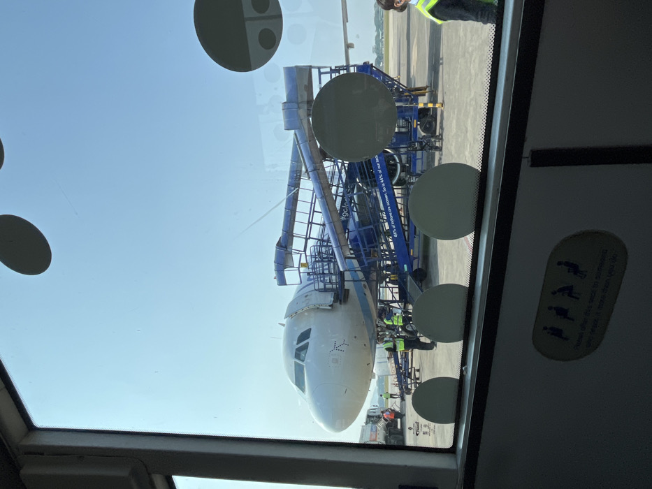 The image, taken from inside a building or vehicle at an airport, shows an airplane parked on the tarmac under a bright, clear sky. The view is partially obstructed by a window, featuring several large circular adhesive pads or suction cups.
The main subject is the front section of a white passenger airplane, likely an Airbus A320 series given the cockpit windows and nose cone shape, which appears to be from IndiGo (a common airline in India, aligning with the Guwahati context). The plane is connected to a blue jet bridge or mobile passenger stairs on its left side. Ground crew members, wearing high-visibility vests, are visible near the aircraft's nose, suggesting pre-flight or post-flight operations. A luggage conveyor belt is also partially visible on the right.
The overall scene depicts standard airport ground operations during daylight hours. The weather appears sunny and clear.
Visible text includes "#gclod" and partially "4% of Indians..." on the blue jet bridge/stairs, likely part of an advertisement or branding. Inside the viewing window, a sticker with pictograms of seated individuals reads "Please offer this seat to someone who needs it more than you do," indicating the observer is within an area with public seating, such as an airport bus or terminal lounge. The location is Guwahati, India.