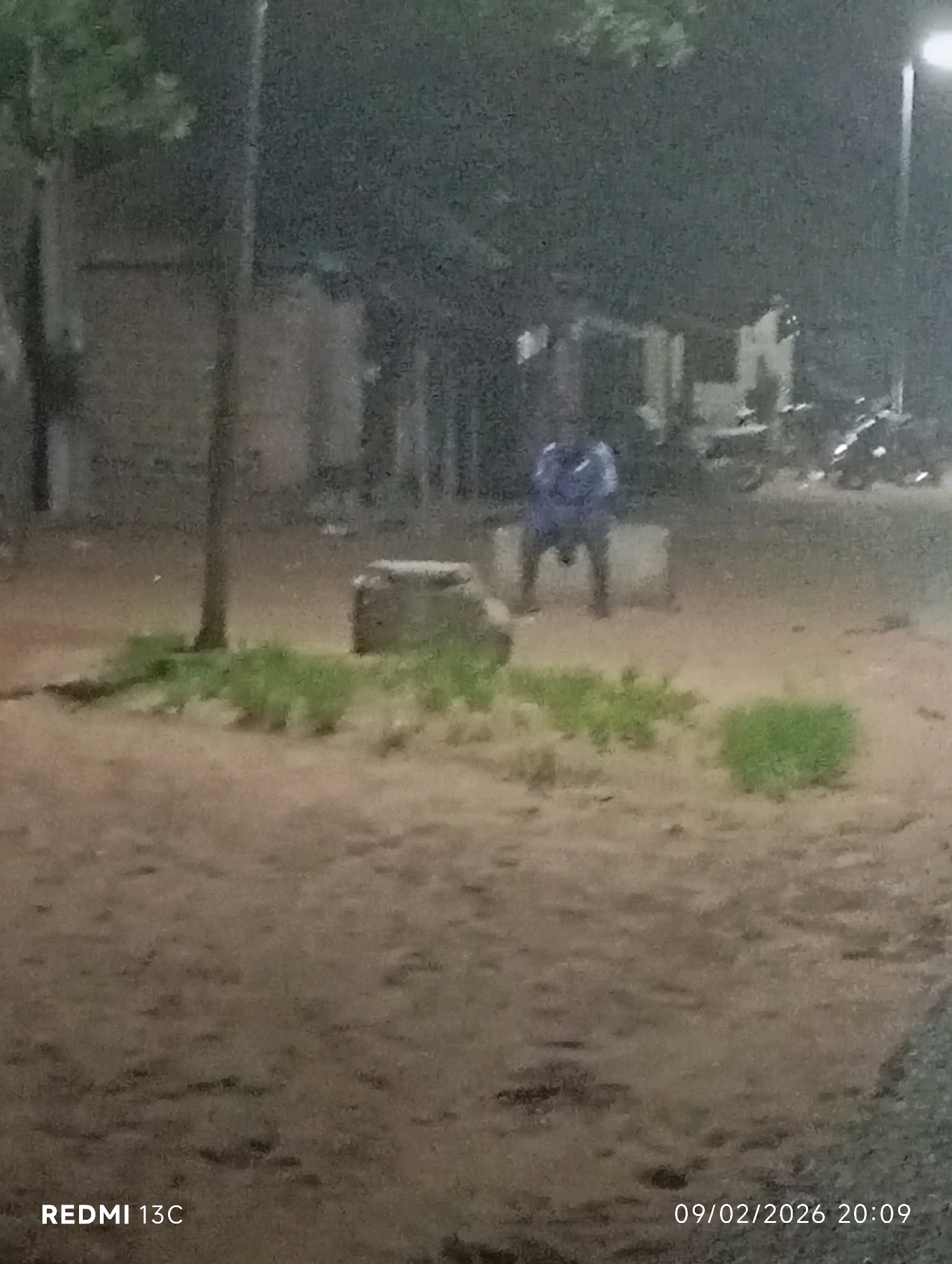 The image captures a nighttime scene, likely in the city of Shendam, Nigeria. A lone individual, clad in a blue garment, is seated or crouching on a low, rectangular object. The environment appears to be an outdoor area, with a dirt or sandy ground surface. To the left, a vertical pole stands, possibly a lamppost or a signpost. In the background, structures are faintly visible, and several motorcycles are parked. A bright light source, likely a streetlamp, illuminates the scene from the upper right. The overall image quality is grainy, suggesting it was taken in low light conditions. A timestamp in the lower right corner indicates "09/02/2026 20:09". The text "REDMI 13C" is present in the lower left corner, indicating the device used to capture the image.