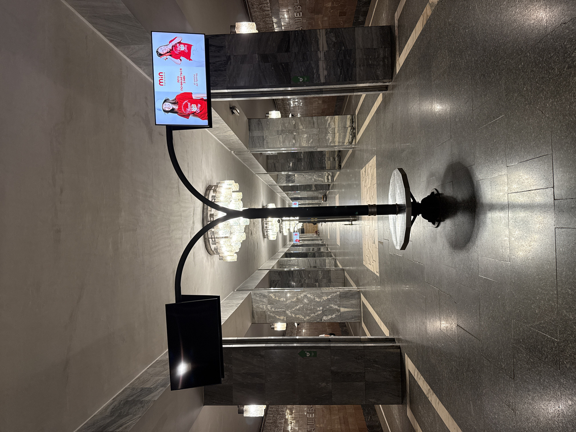 The image depicts the interior of a metro station in Kyiv, Ukraine. It is a long, symmetrical hallway with marble-clad pillars and walls. Two televisions are mounted on a central, arching structure; one displays a black screen, while the other shows an advertisement for a children's clothing brand called "min." The floor is made of dark granite tiles with lighter inlaid geometric patterns. Several elaborate chandeliers hang from the ceiling, casting a warm light. The overall impression is one of Soviet-era architecture, common in many Kyiv metro stations.