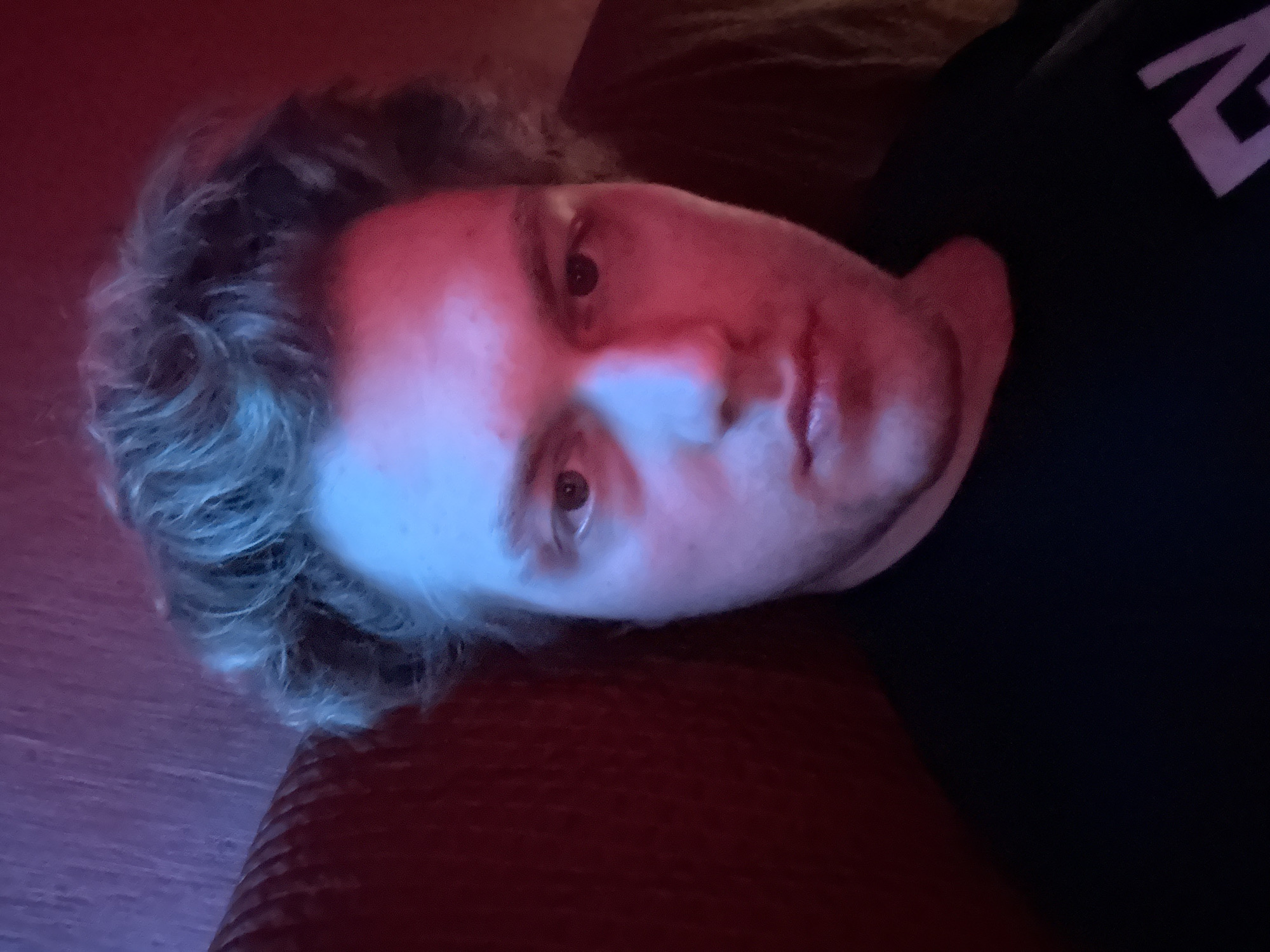 A close-up, overhead, medium shot captures a young person's face bathed in red and blue light. The person is looking directly at the camera with a neutral expression. Their hair is light brown and tousled. They are wearing a black t-shirt with a white graphic on the lower right side of the image. The background is dark, with a red, textured surface on the left, likely a couch or wall. The lighting creates distinct shadows and highlights on the person's face, emphasizing their features. The location is the city of Khimki, Russia.