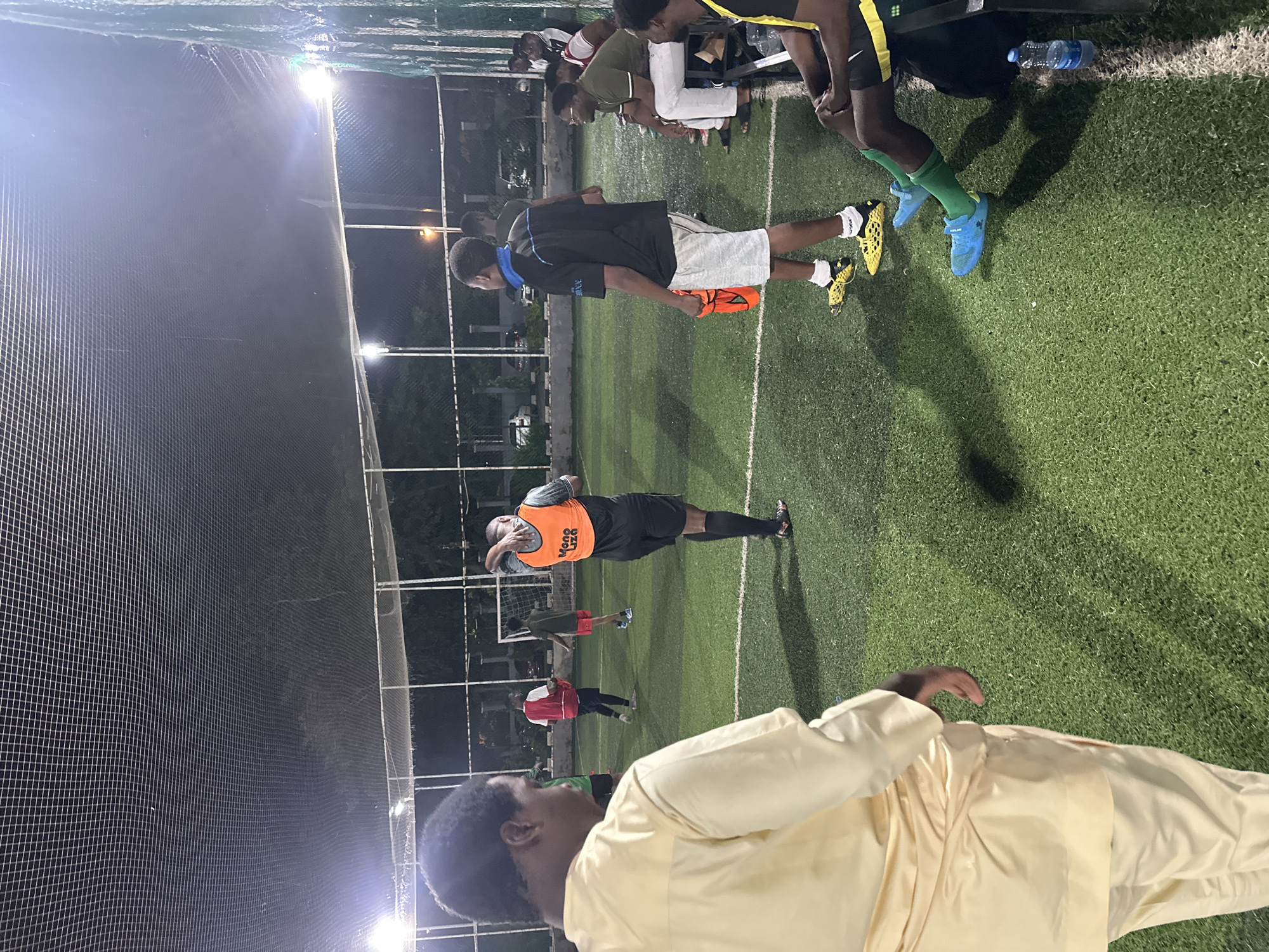 A group of young men are on an artificial turf soccer field at night in Abuja, Nigeria. The field is enclosed by a fence and netting overhead, with stadium lights illuminating the area. The focus is on a man in the foreground wearing a cream-colored traditional outfit, with his back to the camera. Several other men are scattered across the field, some in athletic wear and bibs, suggesting a casual game or practice is taking place. One man is wearing an orange bib with "Mono Liza" printed on it. Others are sitting on a bench on the side of the field. A water bottle is visible near the bench.