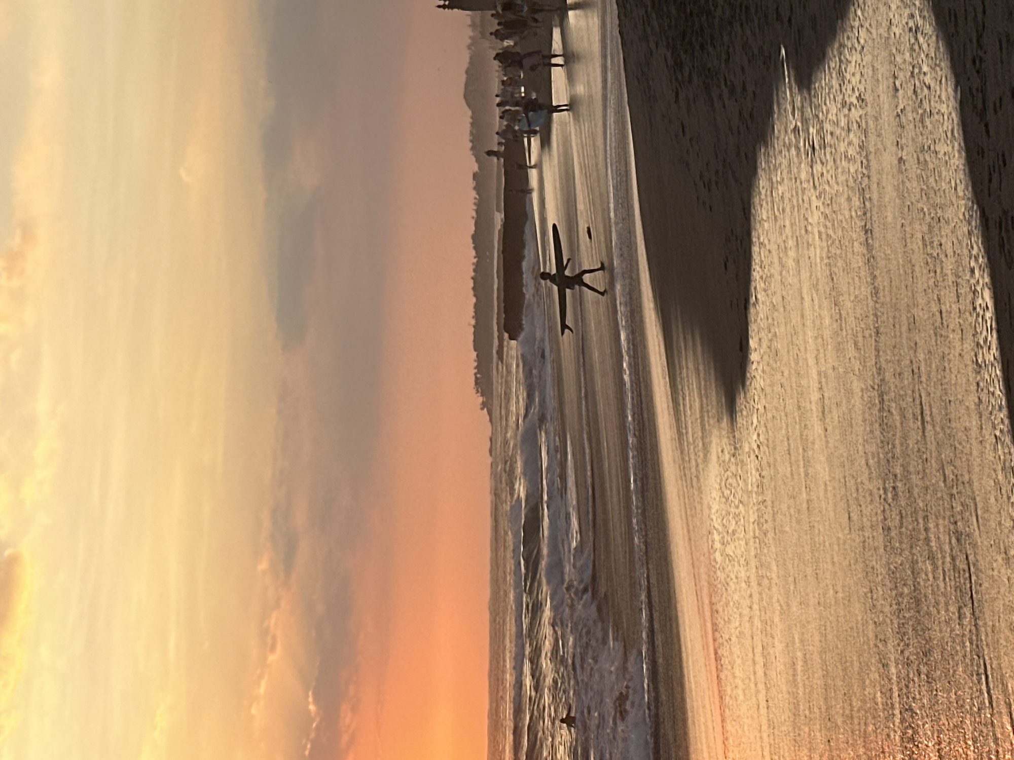 At sunset on a beach in North Kuta, Indonesia, a young person walks along the wet sand carrying a surfboard. The sky is a gradient of orange and yellow, with scattered clouds. Gentle waves roll onto the shore, creating a wet sheen on the sand that reflects the sky's warm hues. In the background, a distant treeline and a few structures are silhouetted against the horizon. Other people are visible further down the beach, some standing near the water and others walking. The overall scene is peaceful and serene, capturing the beauty of a tropical evening.