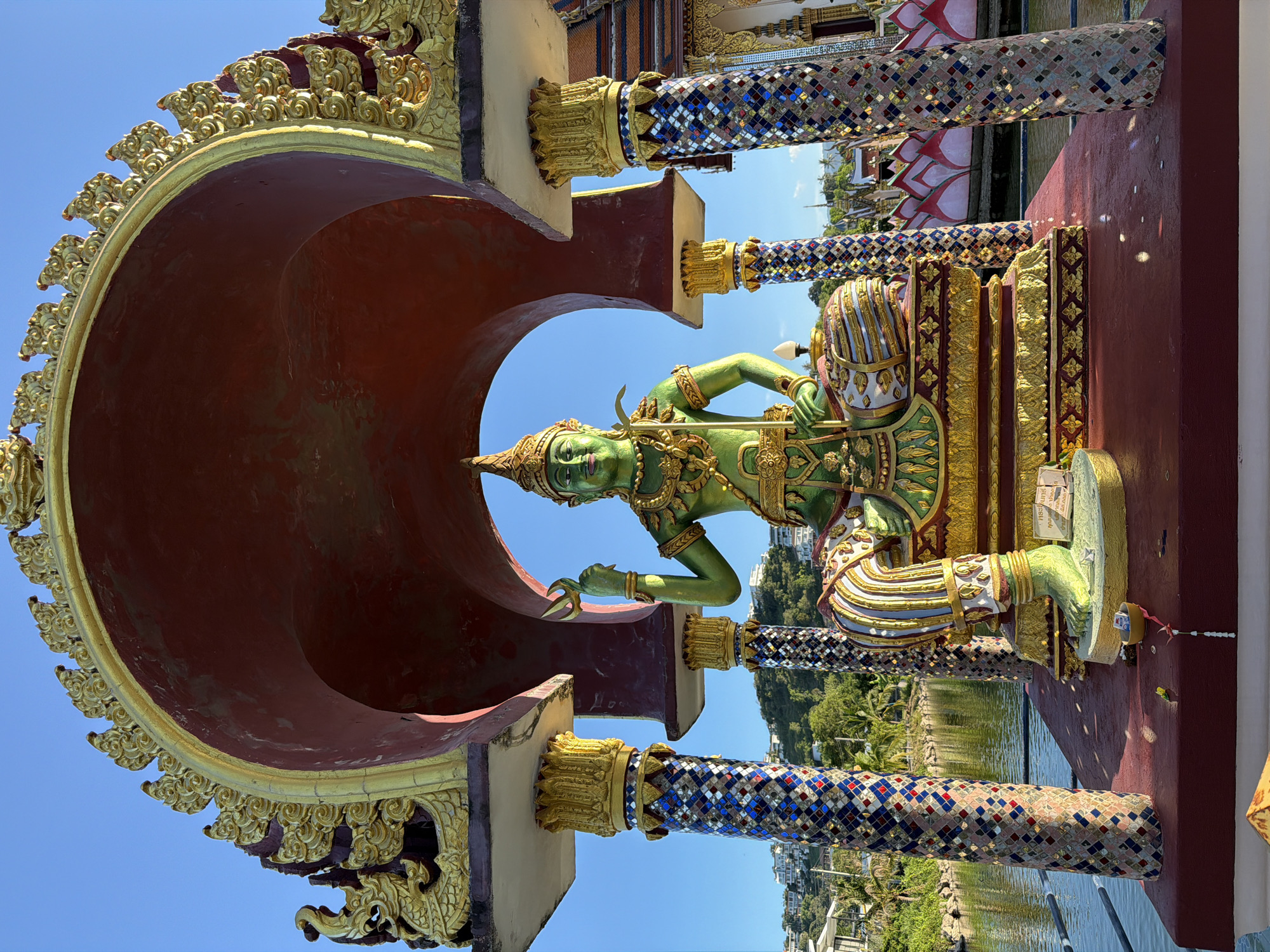 The image captures a vibrant, ornate statue of a green, gilded deity seated within an arched alcove. The deity is adorned with intricate gold jewelry, a conical headdress, and wears patterned lower garments. The statue is positioned on a raised platform in what appears to be a temple complex. The architecture surrounding the statue is rich with detail; the alcove is painted a deep red and trimmed with elaborate gold scrollwork. Supporting this structure are two mosaic-covered pillars, a dazzling display of blue, white, red, and gold tiles.
The statue is situated outdoors, with a body of water, likely a pond or canal, visible in the background. Beyond the water, a lush green hillside is dotted with buildings, suggesting an urban or semi-urban setting. The sky above is a clear, bright blue, indicating fair weather. The overall impression is one of cultural richness and spiritual significance, characteristic of Buddhist temples. The location is identified as Baan Plai Laem, Thailand.