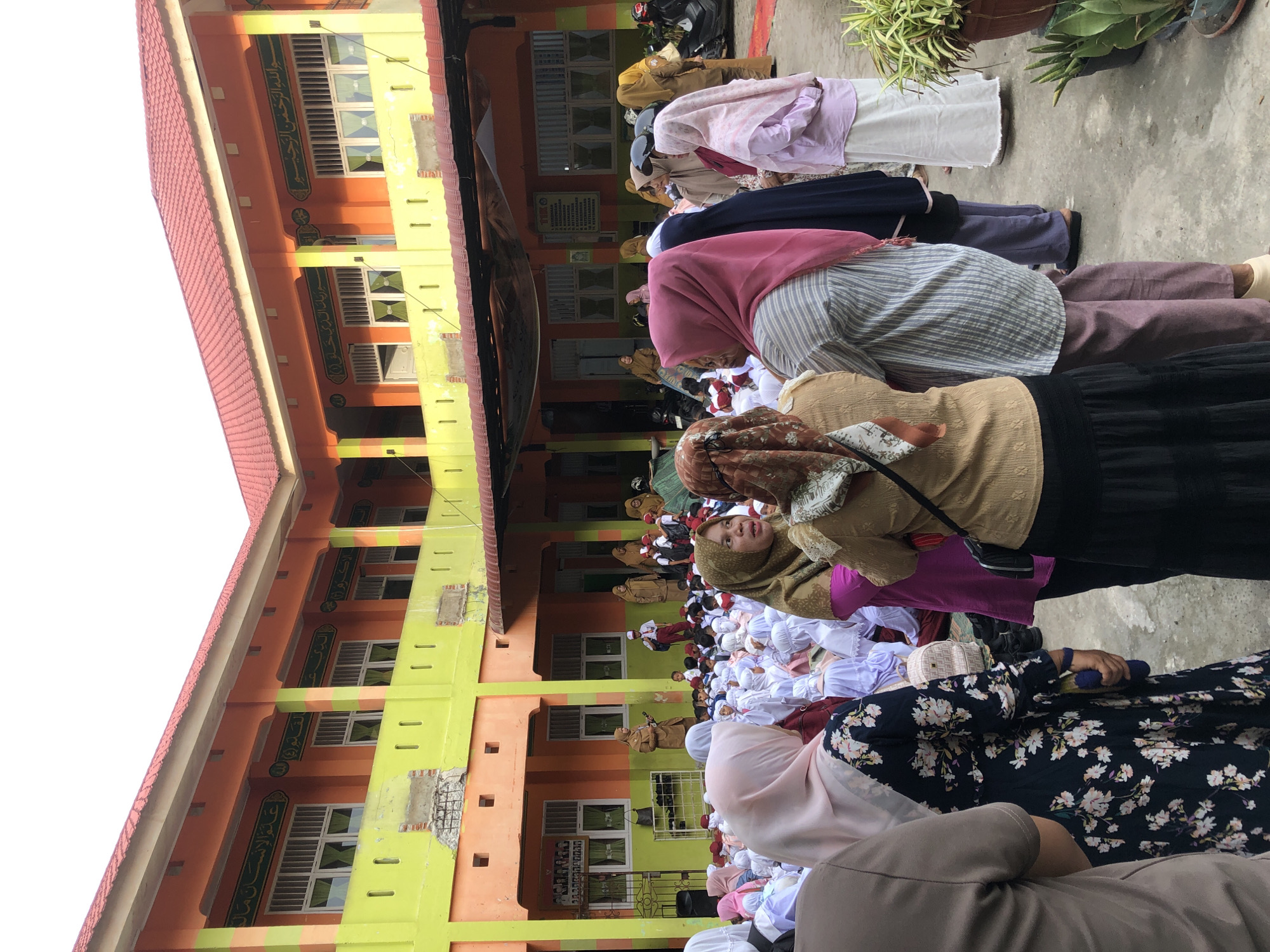 The image shows a gathering of people, mostly women and children, in the courtyard of a school building in Lhokseumawe, Indonesia. The building has an orange and lime green exterior with red roofing. Many of the individuals are wearing hijabs and traditional Islamic attire. Some children are seated on the ground, while others appear to be standing and listening to an adult who is speaking into a microphone. There are motorcycles parked along the side of the courtyard. The scene suggests an outdoor event or assembly taking place at the school.