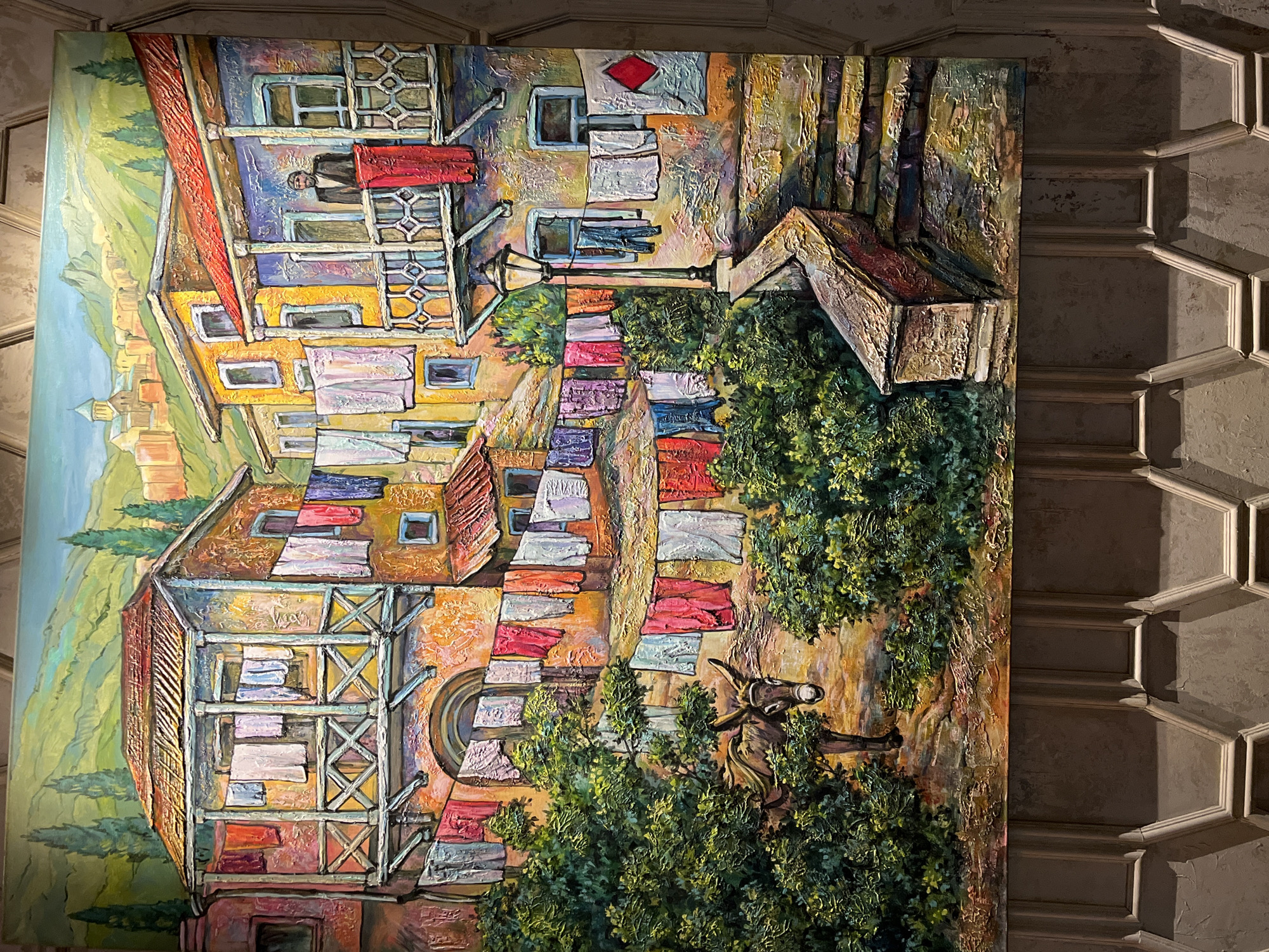 The image is a painting of a narrow European street scene, possibly in Eastern Europe or the Mediterranean, featuring colorful buildings with balconies adorned with laundry hanging out to dry. In the foreground, a donkey stands amidst lush green foliage. A man in a dark suit and hat stands on a balcony in the background, looking out over the street. The architecture suggests an older city with traditional houses. The painting is rendered in a thick, impasto style, giving it a textured, almost sculptural quality. The setting is mountainous with trees and a building with a dome visible in the distance. The painting is mounted on a wall with decorative paneling below. There is no specific indication of the city of Khimki, Russia, within the artwork itself.