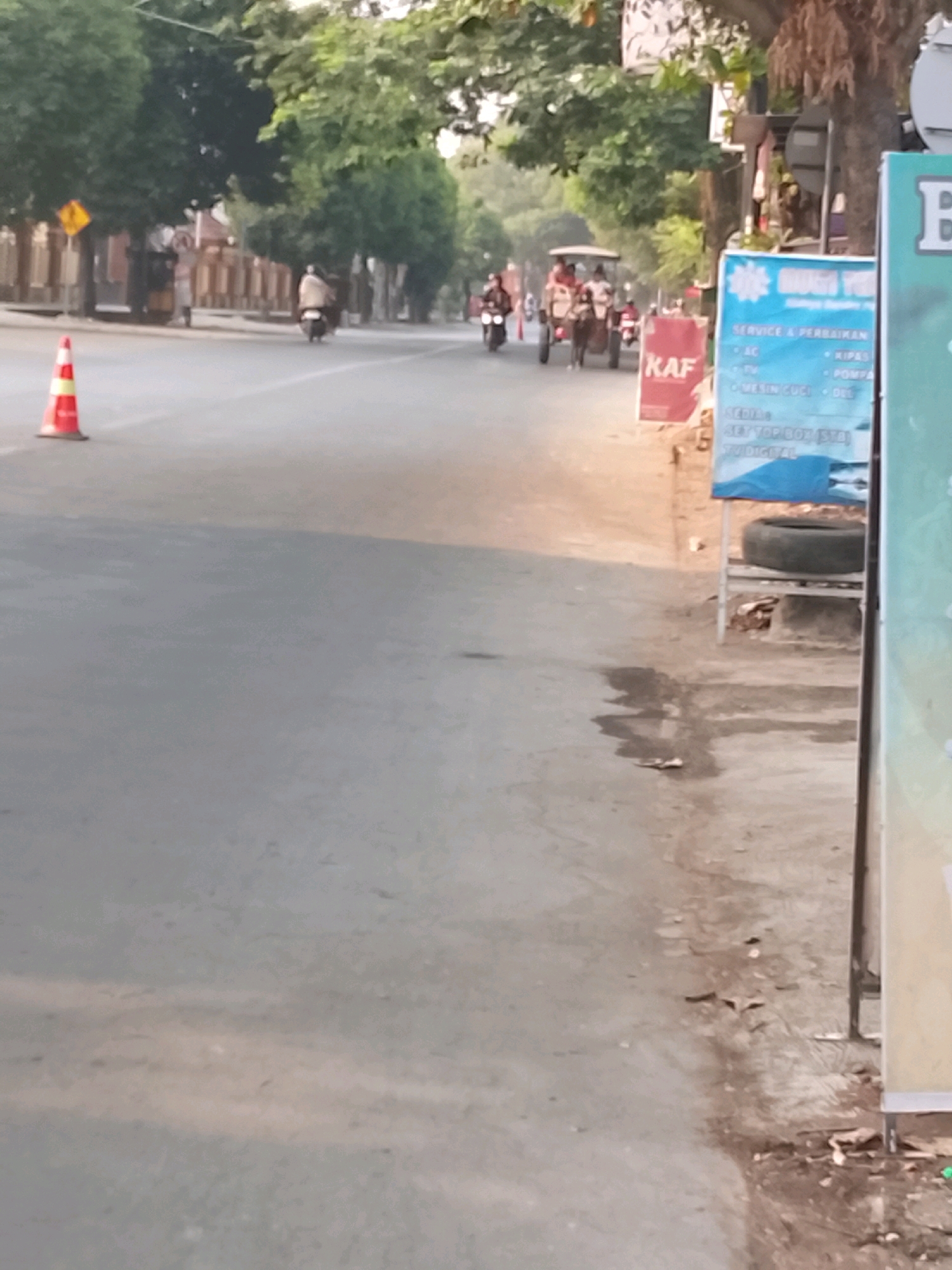 The image captures a street scene in Slawi, Indonesia, possibly along a moderately busy road. On the left side of the road, a bright orange traffic cone stands on the asphalt, suggesting potential road work or a hazard. Beyond the cone, a yellow diamond-shaped road sign is visible. Further down the road, a person rides a motorcycle, their form obscured by a blanket or shawl.
In the center of the road, a horse-drawn cart is in motion, carrying multiple passengers. Several other motorcycles are also present on the road, some with riders. The street is lined with trees on the left, their foliage creating a dappled shade.
On the right side of the image, a blue and white sign board is displayed, listing various services such as "SERVICE & PERBAIKAN," "AC," "TV," "MESIN CUCI," and "MEDIA SET TOP BOX (STB) TV DIGITAL." Next to this sign, a spare tire is placed on a metal shelf, with some debris and dirt on the ground. Another red advertisement board with the text "KAF" is also visible. The overall environment appears to be a typical urban or semi-urban street in Indonesia, with evidence of both modern and traditional modes of transportation. The lighting suggests it might be late afternoon or early morning.