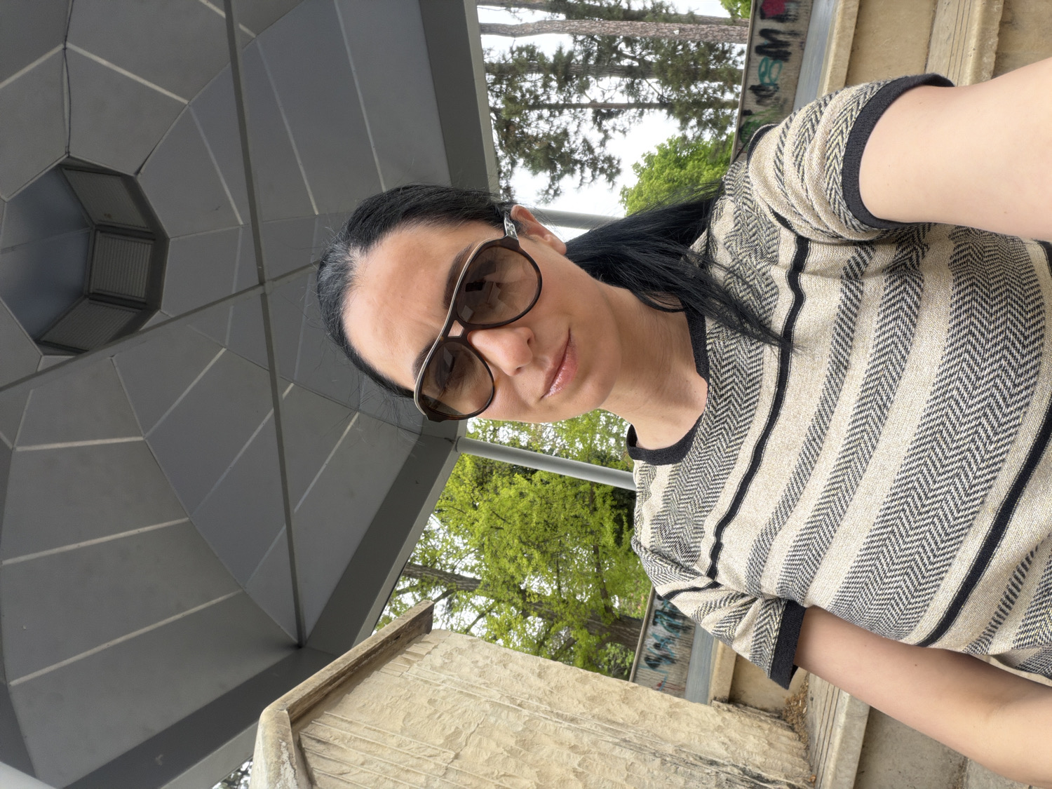 A mid-shot selfie features a woman posing outdoors in Pleven, Bulgaria, looking directly at the camera with a slight smile. She wears large, brown-tinted sunglasses with dark frames and a short-sleeved top characterized by alternating horizontal stripes: some solid dark, others a textured herringbone pattern in light and dark tones. Her dark hair is pulled back.
The image is captured from a low, upward-looking angle and is notably tilted. In the upper left of the frame, a prominent dark grey, multi-faceted geometric structure, possibly part of a modern pavilion roof or overhang, features a light fixture embedded within its design. To the woman's right and extending into the lower background, a light-colored, textured stone wall or barrier is visible. Behind her, a backdrop of trees with vibrant green foliage suggests a spring or summer day, with some bare tree trunks also in view. Patches of bright, possibly overcast, sky are visible, confirming it is daytime. On a distant wall in the upper right background, some indistinct graffiti can be seen. The setting evokes a public park or a modern architectural space within Pleven.