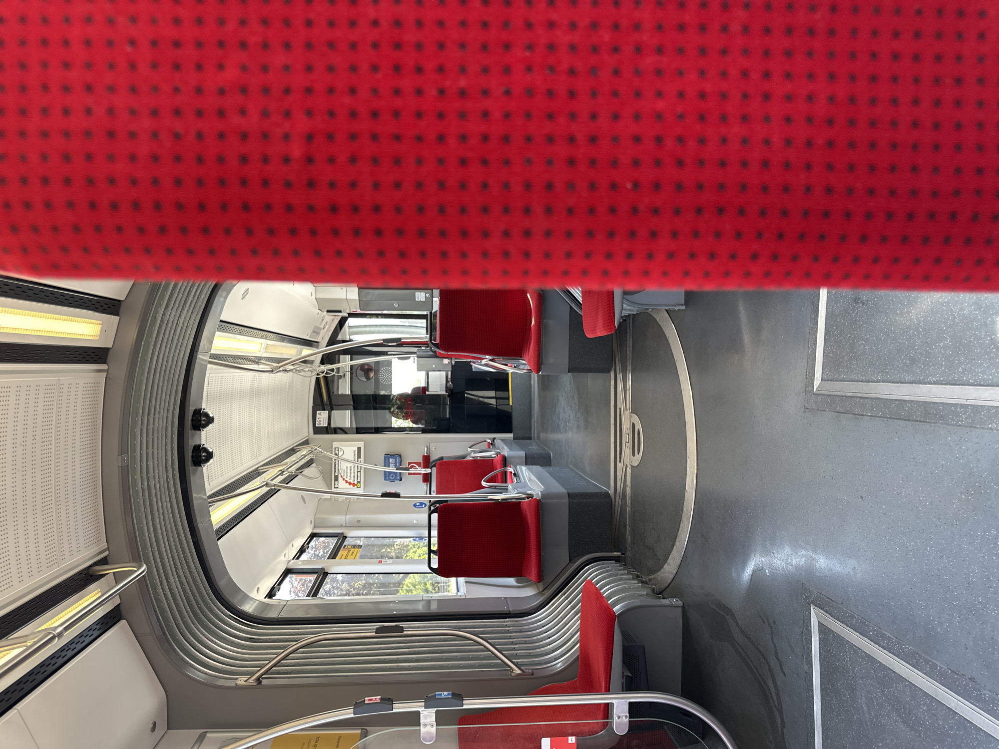 The image shows the interior of a modern tram or light rail vehicle in Krefeld, Germany. The tram has red upholstered seats with a black dotted pattern on the seat backs and seat cushions visible. The seats are arranged in rows on both sides of the aisle, with some seats facing forward and others backward. The floor is a gray speckled material with metallic trim around designated standing areas. The ceiling features fluorescent lighting and metal handrails. A person is visible standing near the front of the tram, wearing a dark jacket and a red hooded garment. The tram appears to be stationary, with no visible passengers seated. The overall environment is clean and well-lit, suggesting a functional public transport vehicle.