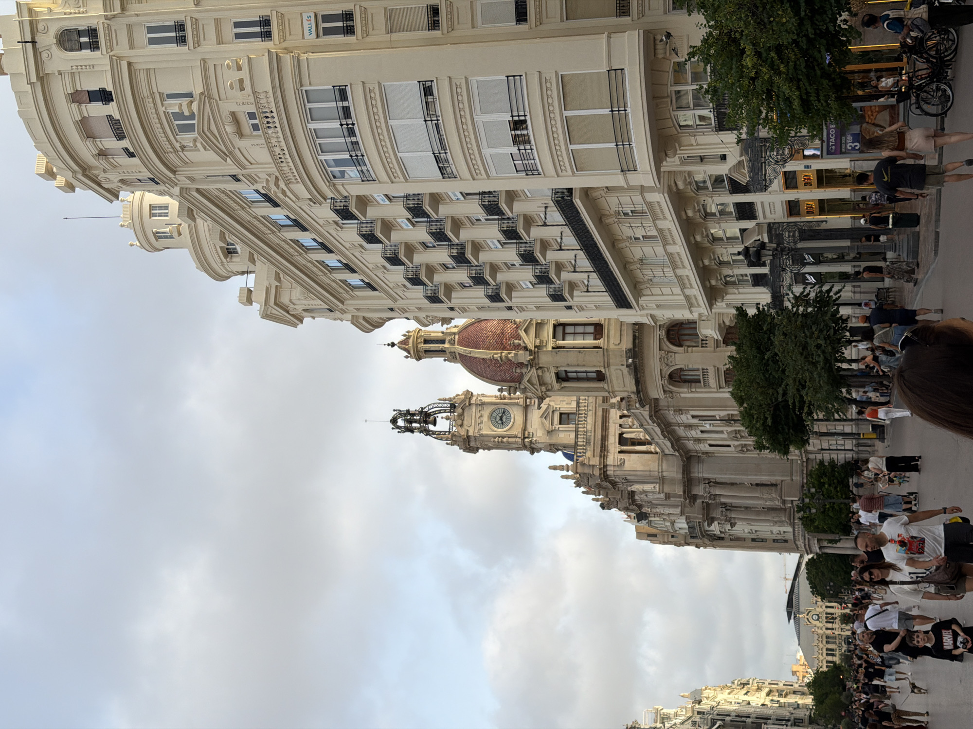 The image depicts a street scene in a European city, likely Valencia, Spain, based on the architecture and signage. In the foreground, a crowd of people walks down a wide, paved street lined with ornate, cream-colored buildings. The buildings feature balconies, large windows, and classical architectural elements, with one prominent building showcasing a red-domed roof and a clock tower. Several shops and cafes are visible along the street level, with some signage in Spanish. A few trees are planted along the sidewalk, and a lamppost stands near the entrance of a shop. In the background, more buildings and a distant structure with a large roof are visible. The sky is cloudy, suggesting it might be late afternoon or early evening. People are dressed in casual summer attire. Some individuals are walking, while others are standing or conversing. One person is walking a small dog. The overall impression is of a busy urban environment.