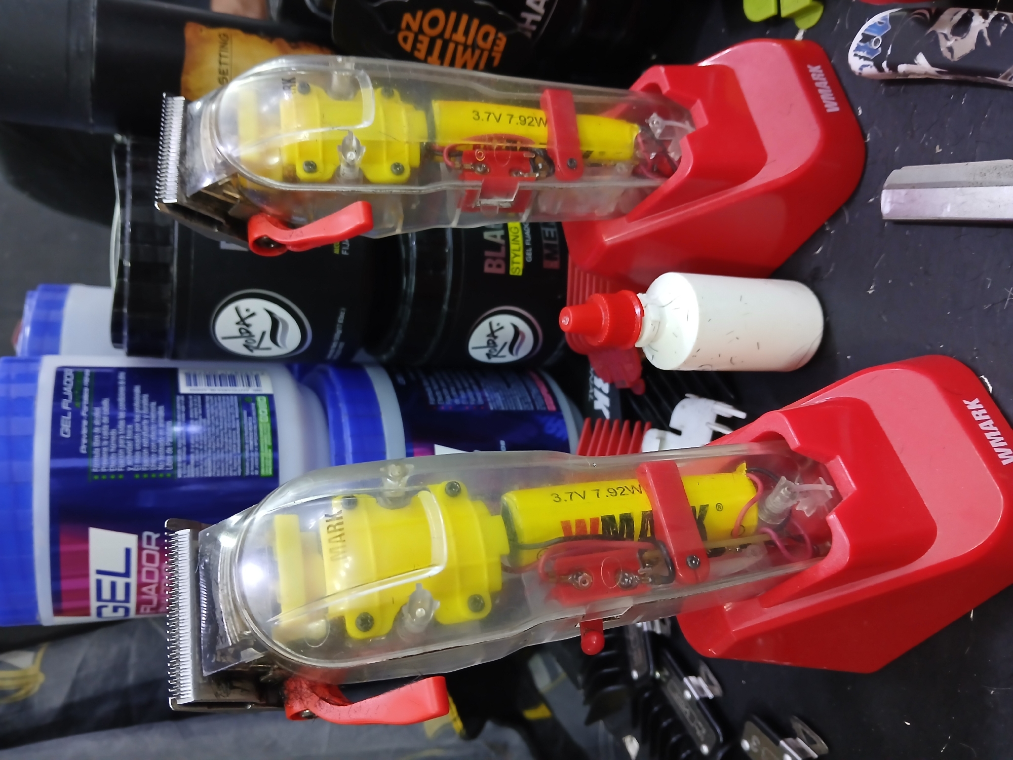 The image shows two Wmark brand hair clippers, each with a transparent body revealing a yellow battery (3.7V 7.92W) and motor, and a red plastic lever and charging stand. The clippers are placed on a dark surface alongside various barbering products including containers labeled "GEL FIJADOR" and "Roda Styling Gel". A white bottle with a red nozzle and a straight razor are also visible. The specific location in Juan Griego, Venezuela cannot be determined from the image.