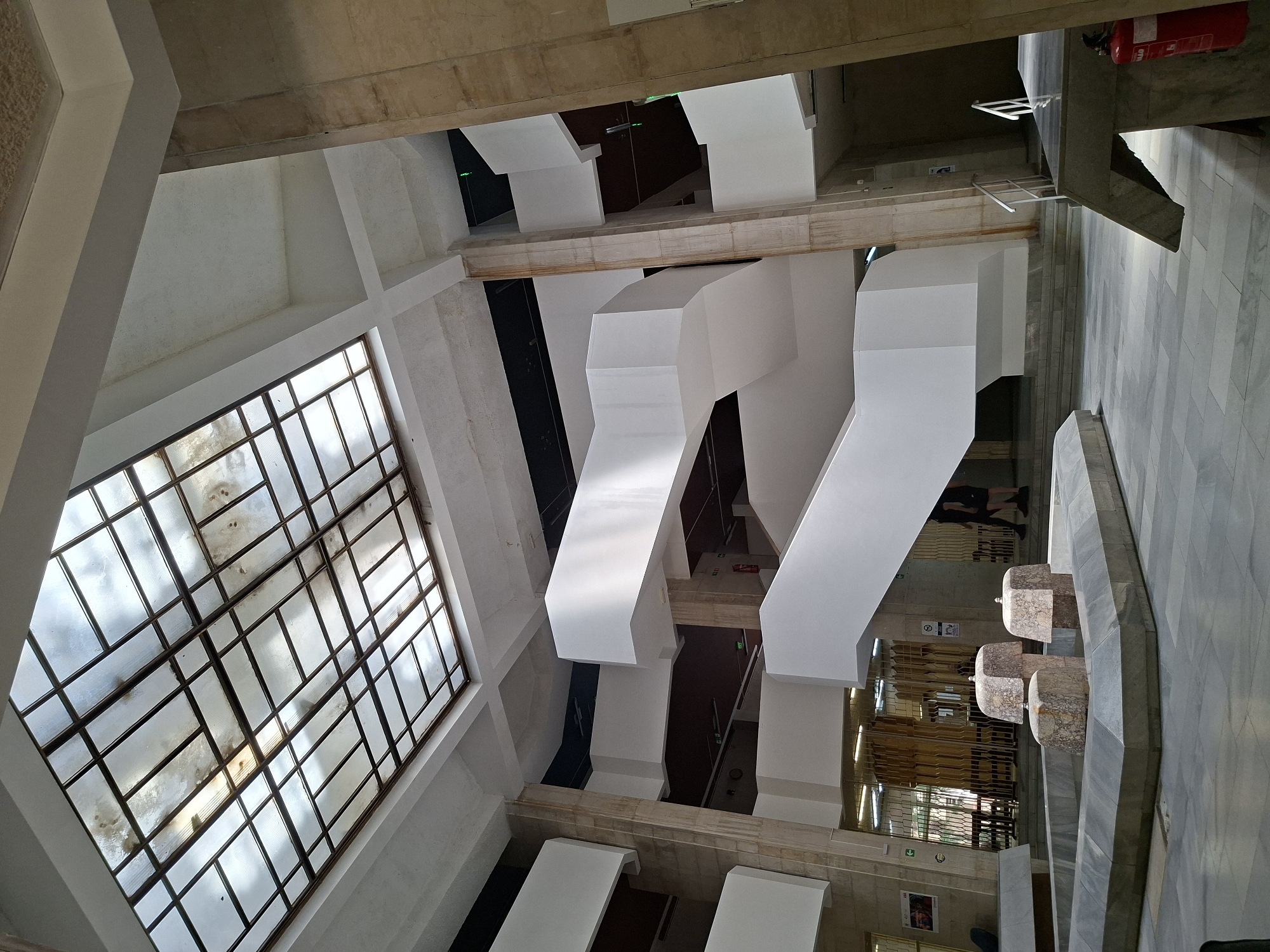 This media file presents a high-angle view looking down into a multi-level indoor architectural space, likely a public building or complex in Plovdiv, Bulgaria. The perspective is rotated clockwise, creating dynamic diagonal lines throughout the composition.
On the left side, a large, multi-paned window or skylight with a grid pattern dominates the wall, allowing ample natural light into the space. The window glass appears somewhat aged or dirty. Across the center and extending to the right, striking white, angular, and geometric structures project into the open area, possibly functioning as ventilation ducts, light fixtures, or purely decorative architectural elements.
The floor on the right side is covered with large, light-colored tiles, exhibiting a patterned design reminiscent of marble or polished stone. Along this tiled floor, four distinct square-shaped pedestals or decorative elements, crafted from light-colored, speckled stone with rounded tops, are arranged in a line. A red fire extinguisher is mounted on a wall further back on the right.
In the background, darker openings and passages are visible, suggesting lower levels or adjacent rooms. A vague, partial reflection or figure of a person can be discerned through one of these darker areas. Visible text and symbols include a sign with a downward arrow, a circular symbol (possibly a "no" or informational sign), and some partially obscured text on the fire extinguisher, which includes "FIRE."
The scene is static, with no specific activity taking place, conveying a sense of quiet and the architectural details of the interior during what appears to be daytime, indicated by the natural light from the large window.