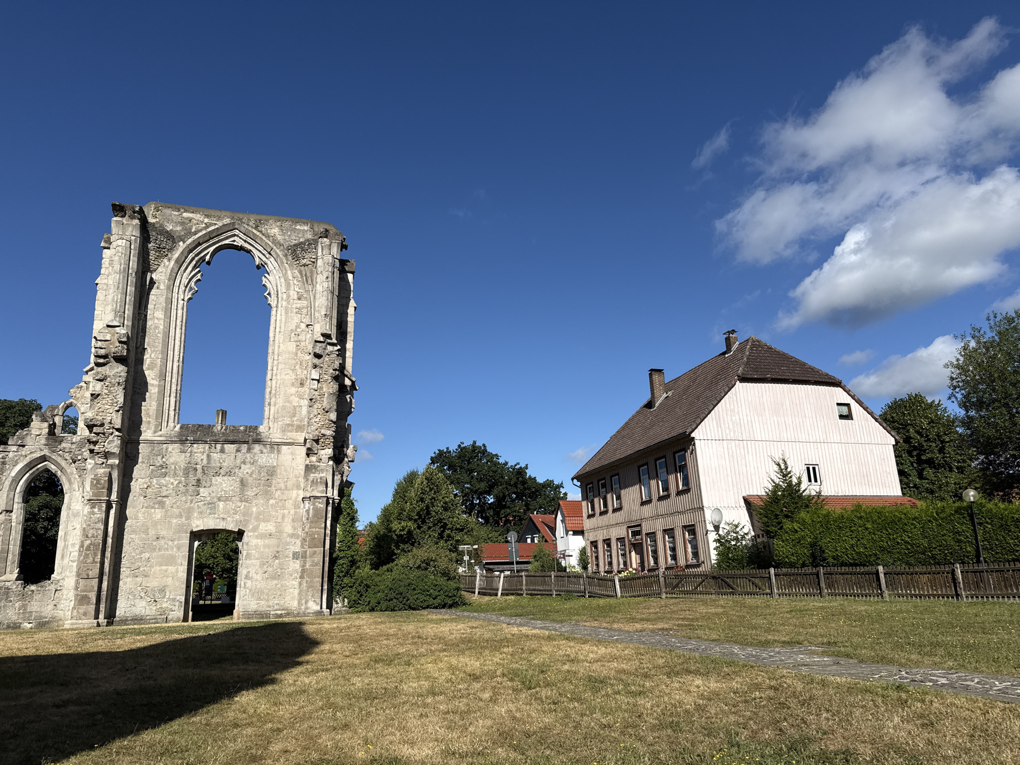 The image depicts the ruins of a Gothic church on the left, with a large arched window opening framing a clear blue sky. In the center and right of the frame, a light-colored, two-story building with a brown tiled roof stands adjacent to the ruins. This building has multiple windows with dark frames and wooden siding on its upper portion. A wooden fence runs along the property line, separating the grassy area with a stone path from the buildings and vegetation. Lush green trees and bushes are visible behind the fence, and scattered white clouds dot the sky. The scene appears to be located in a park-like setting on a sunny day. This is a view of the former Walkenried Abbey, a historic monastery located in Walkenried, Germany.